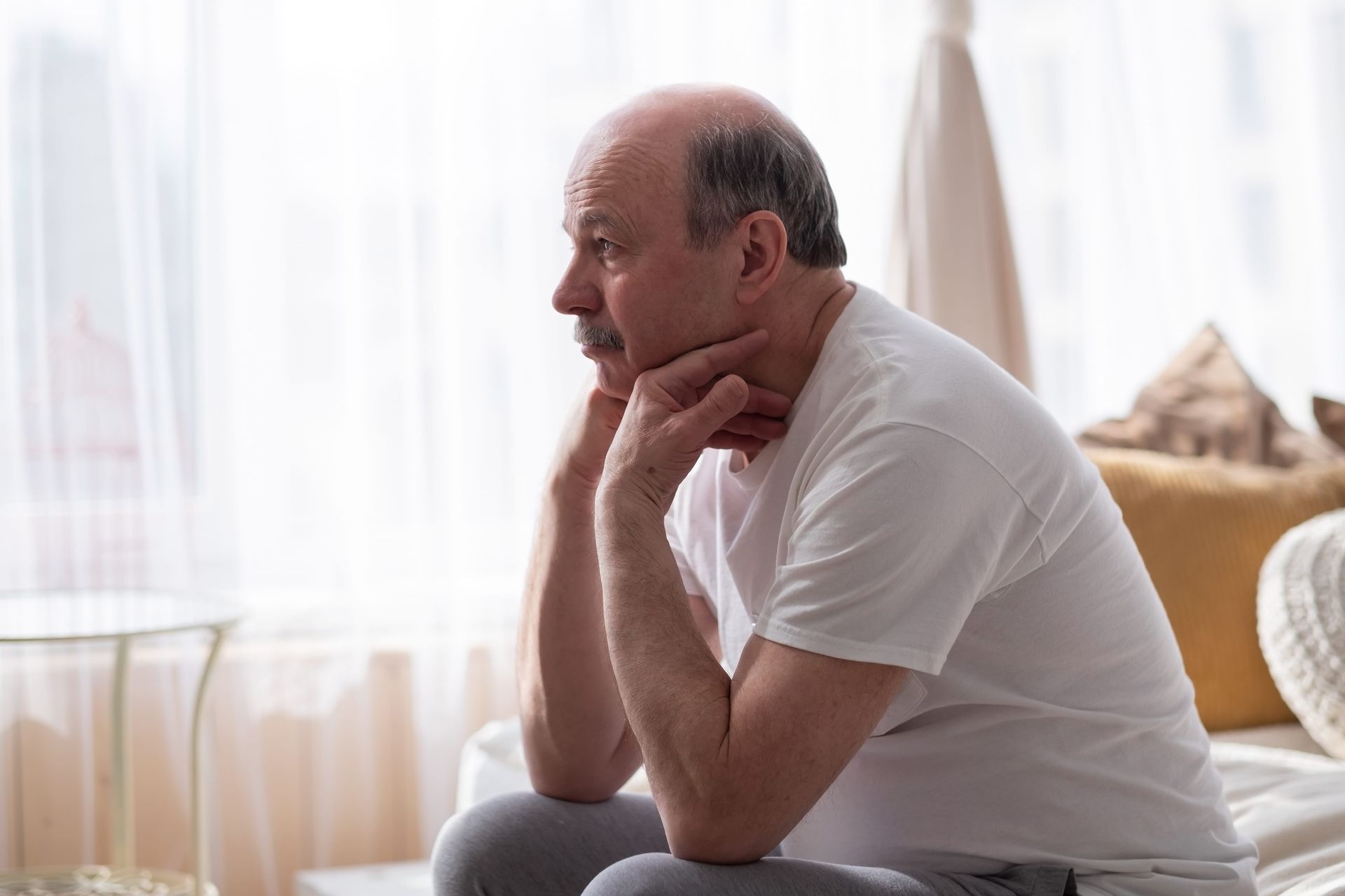 Man sitting on a couch, resting his chin on his hands in a bright living room.