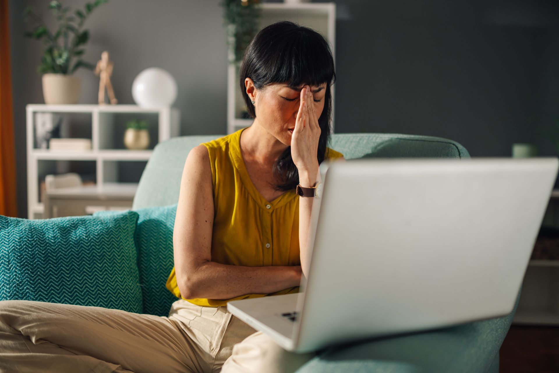 Frustrated person sitting on a couch, covering their face beside an open laptop.