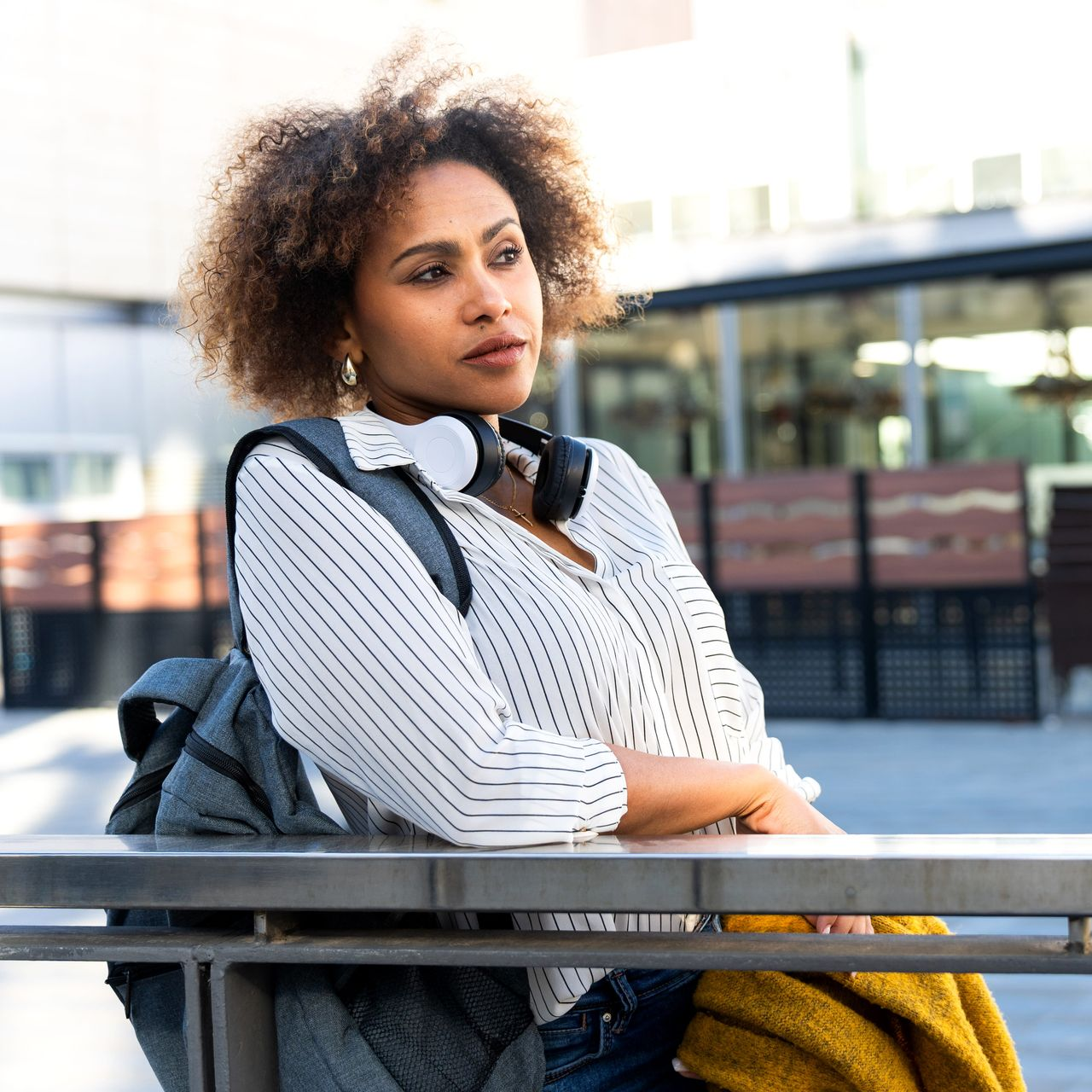 Person leaning on a railing outdoors with headphones around their neck and a backpack nearby