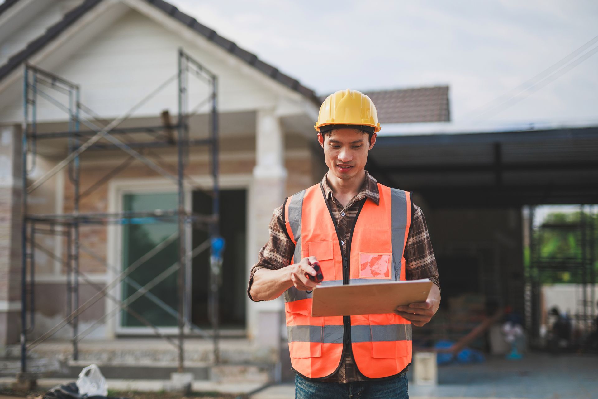 Construction worker in orange vest and hard hat holding a clipboard outside a house under construction