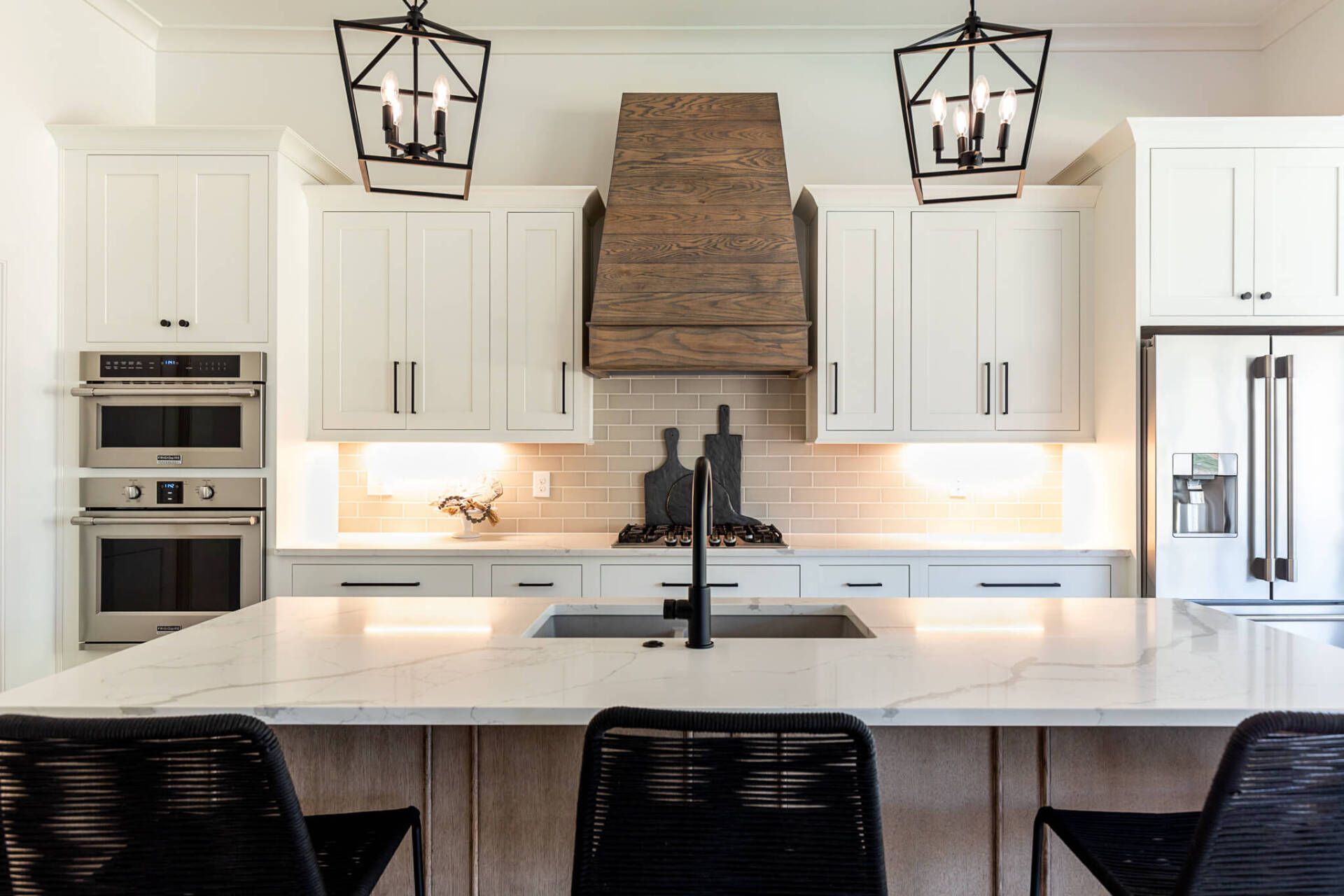 Modern white kitchen with large island, black barstools, built-in ovens, and rustic wood range hood