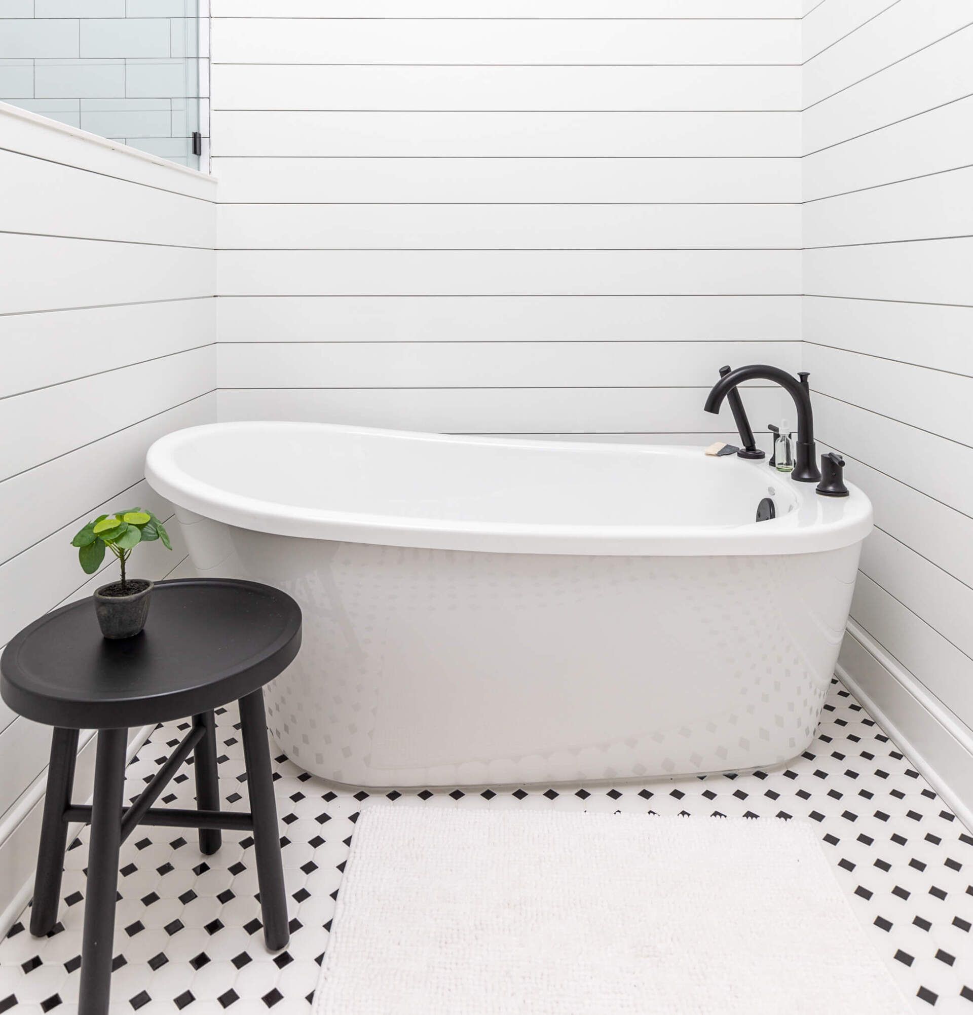 Modern white bathroom with a freestanding tub, black fixtures, stool, and small plant on a black-and-white tiled floor