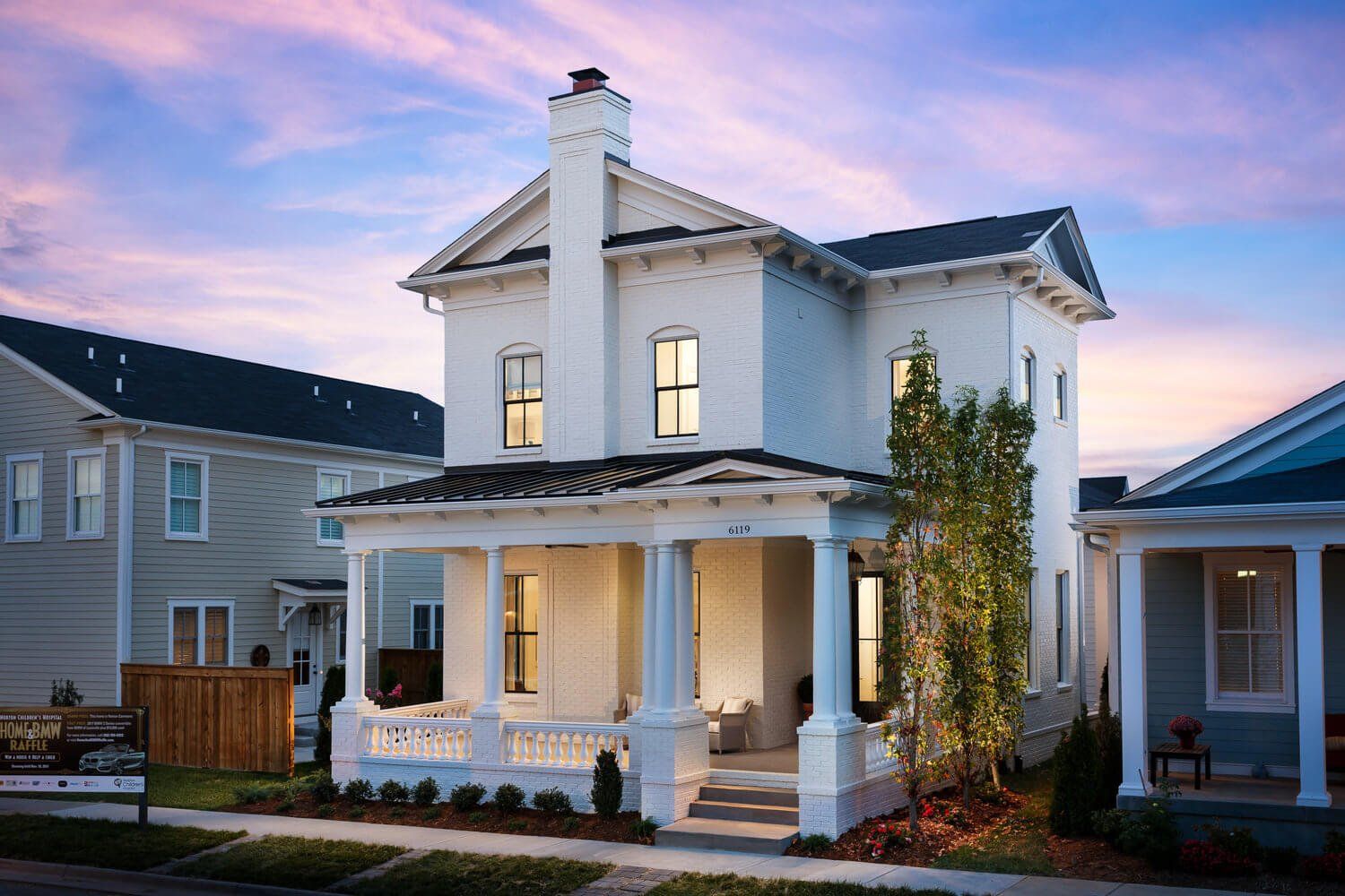 White two-story house with porch at sunset, lit windows, and a pastel sky