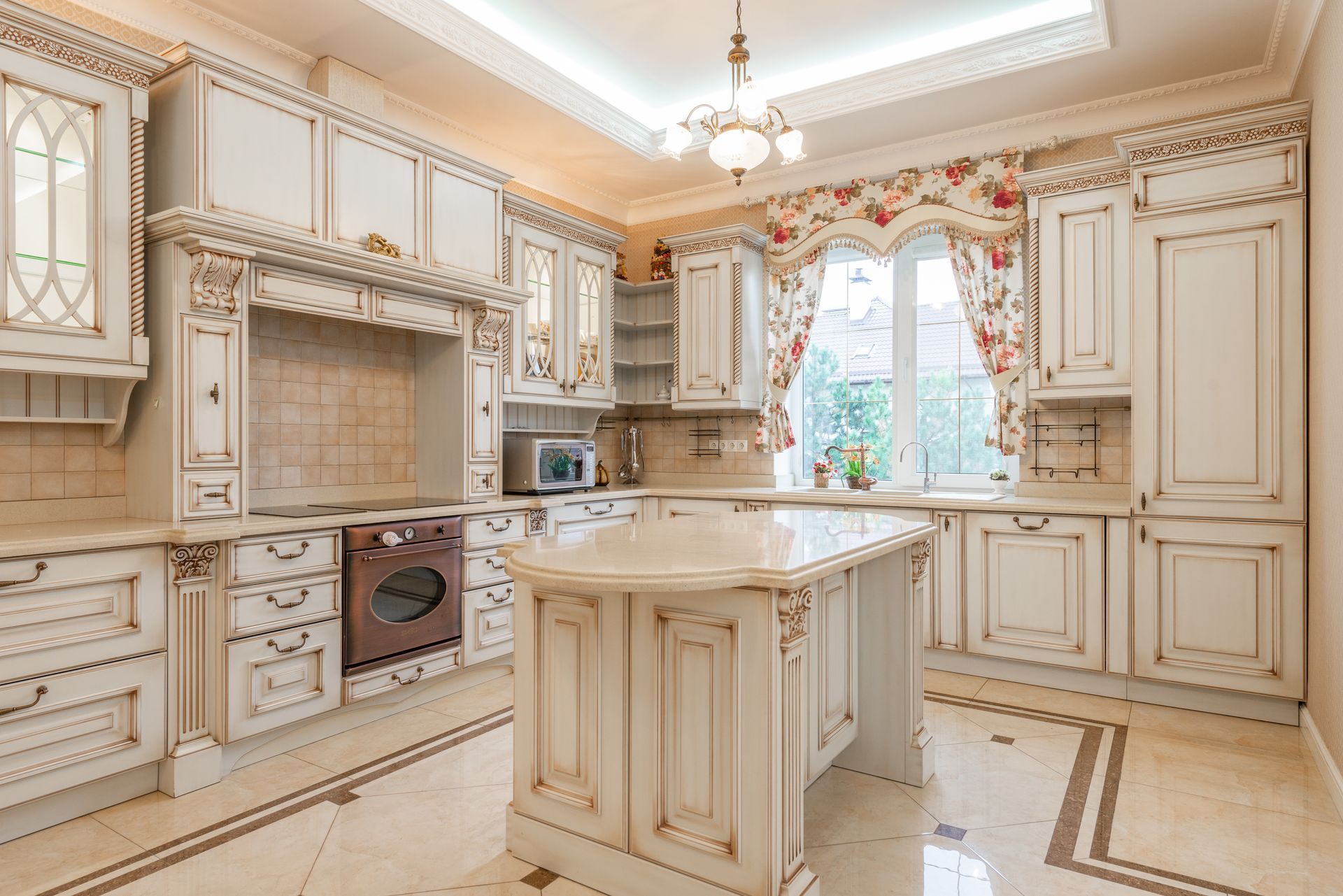 Elegant cream-colored kitchen with ornate cabinets, central island, and window with floral curtains