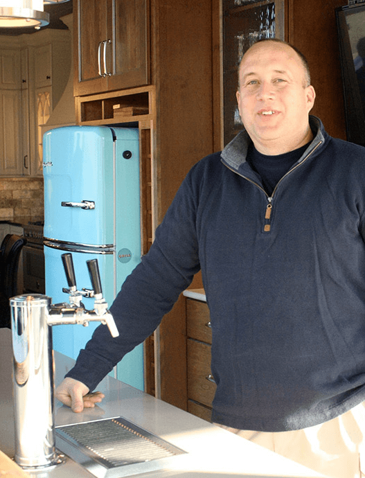 Man standing beside a shiny tap in a kitchen with a blue retro refrigerator and wooden cabinets.