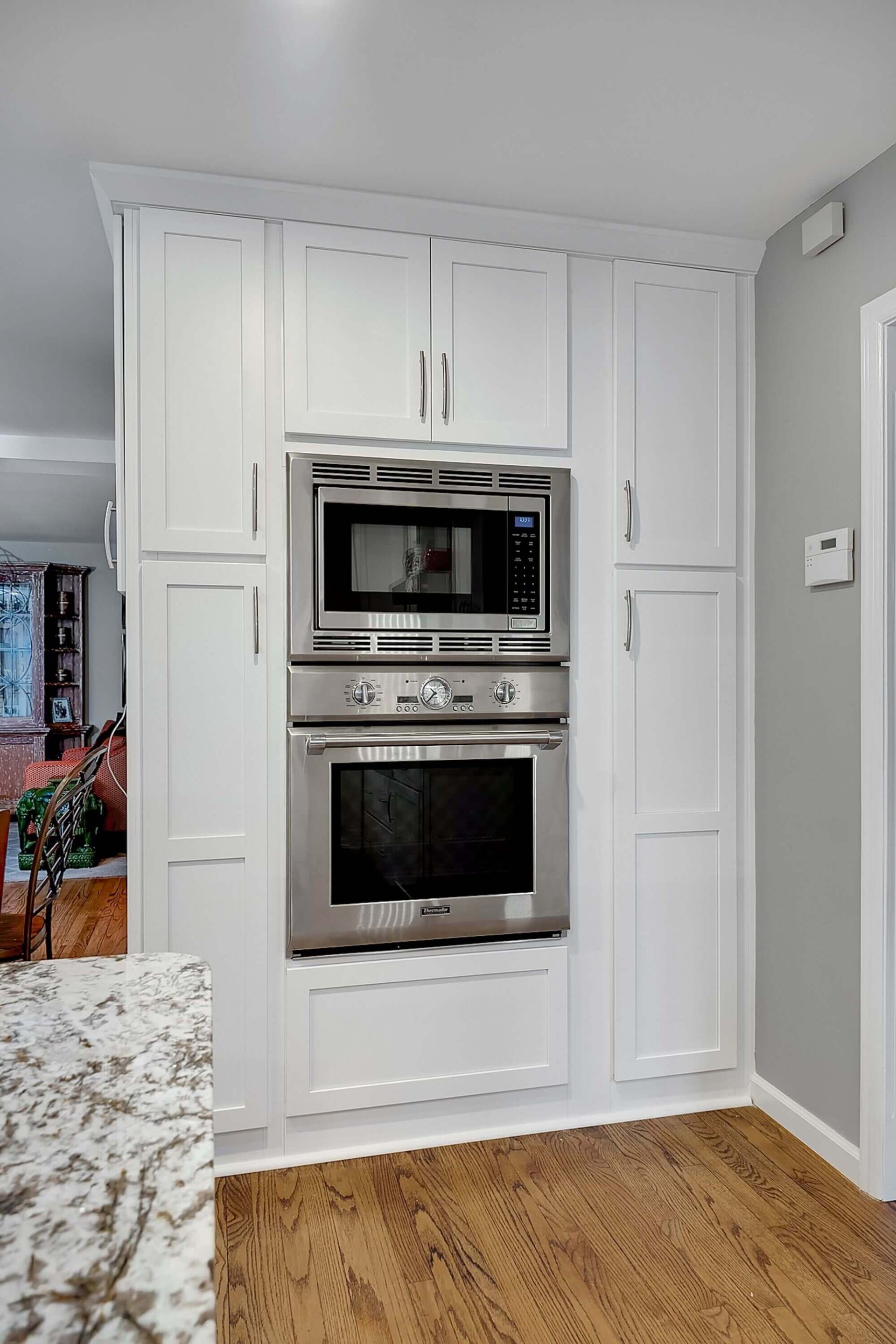 White built-in kitchen cabinet with stacked microwave and oven in a gray room, with wood flooring.