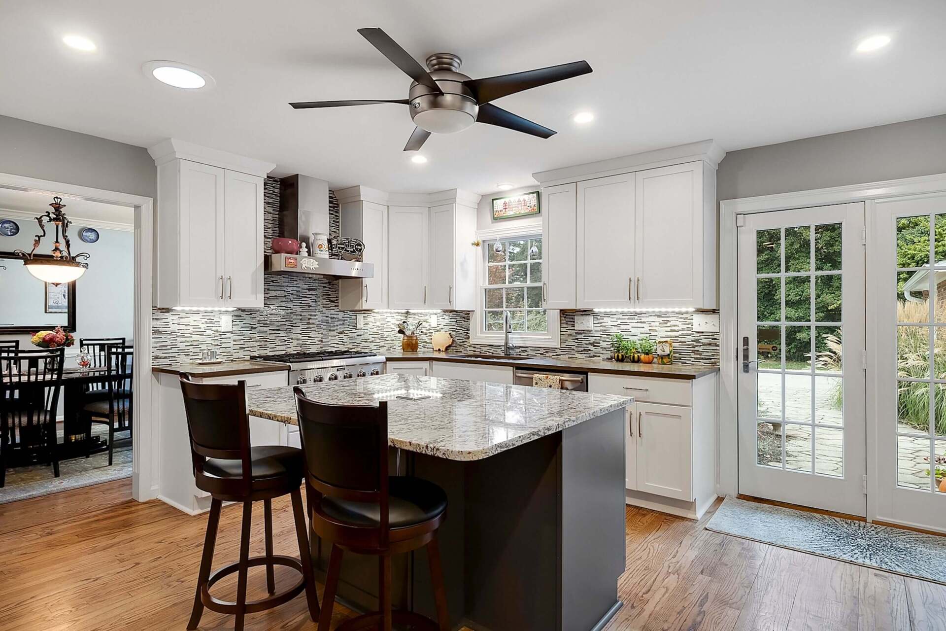 Bright modern kitchen with granite island, white cabinets, bar stools, and French doors to a patio.