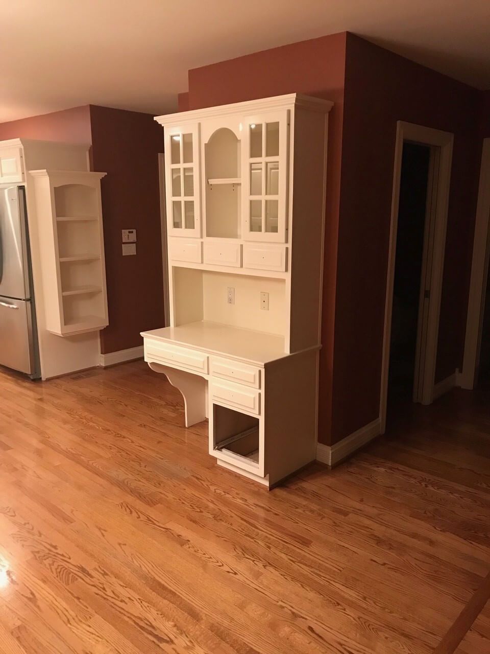 Empty kitchen with hardwood floors, white cabinets, and a red accent wall, viewed from a doorway.