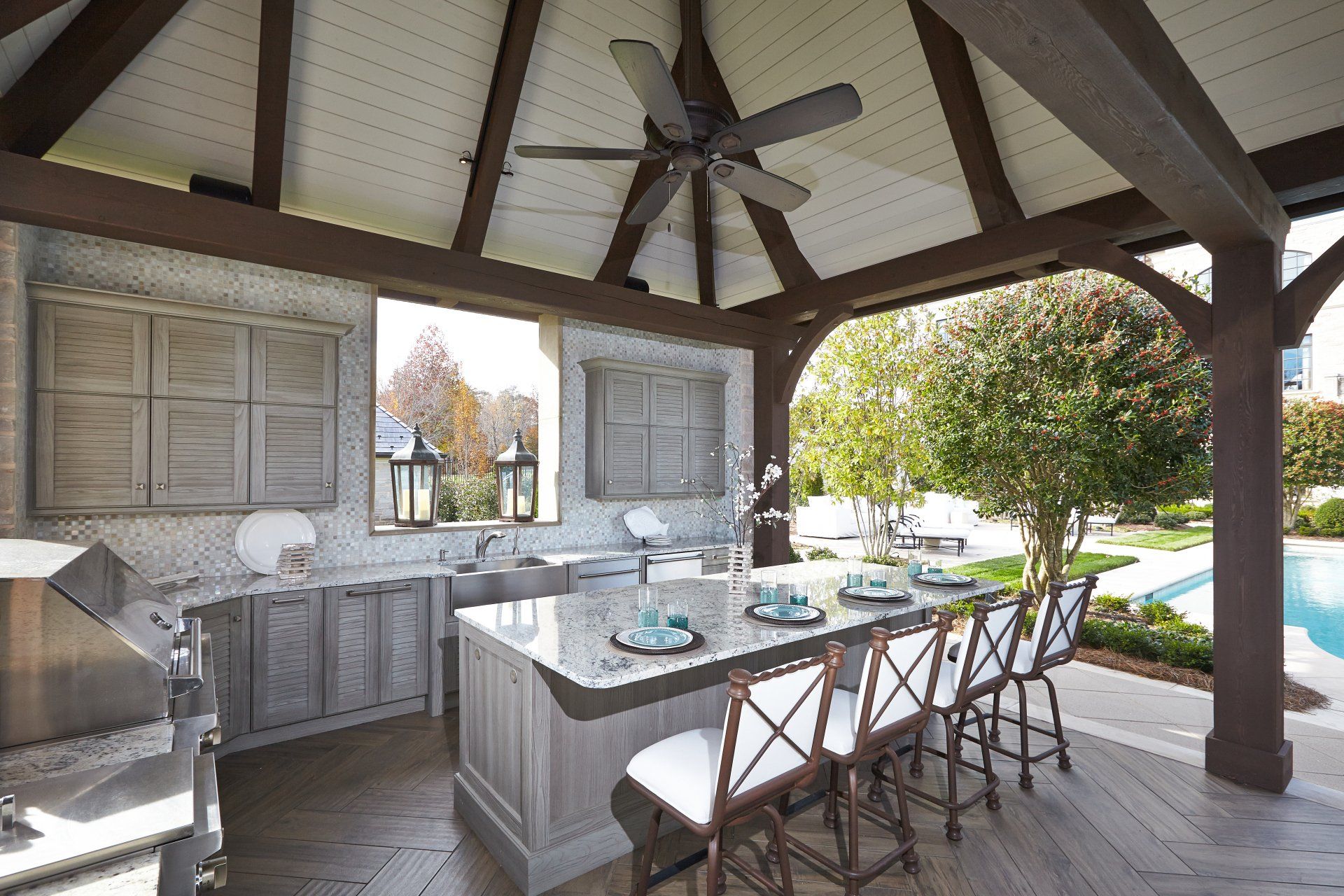Open-air outdoor kitchen and dining area with island, bar stools, wood beams, and pool view