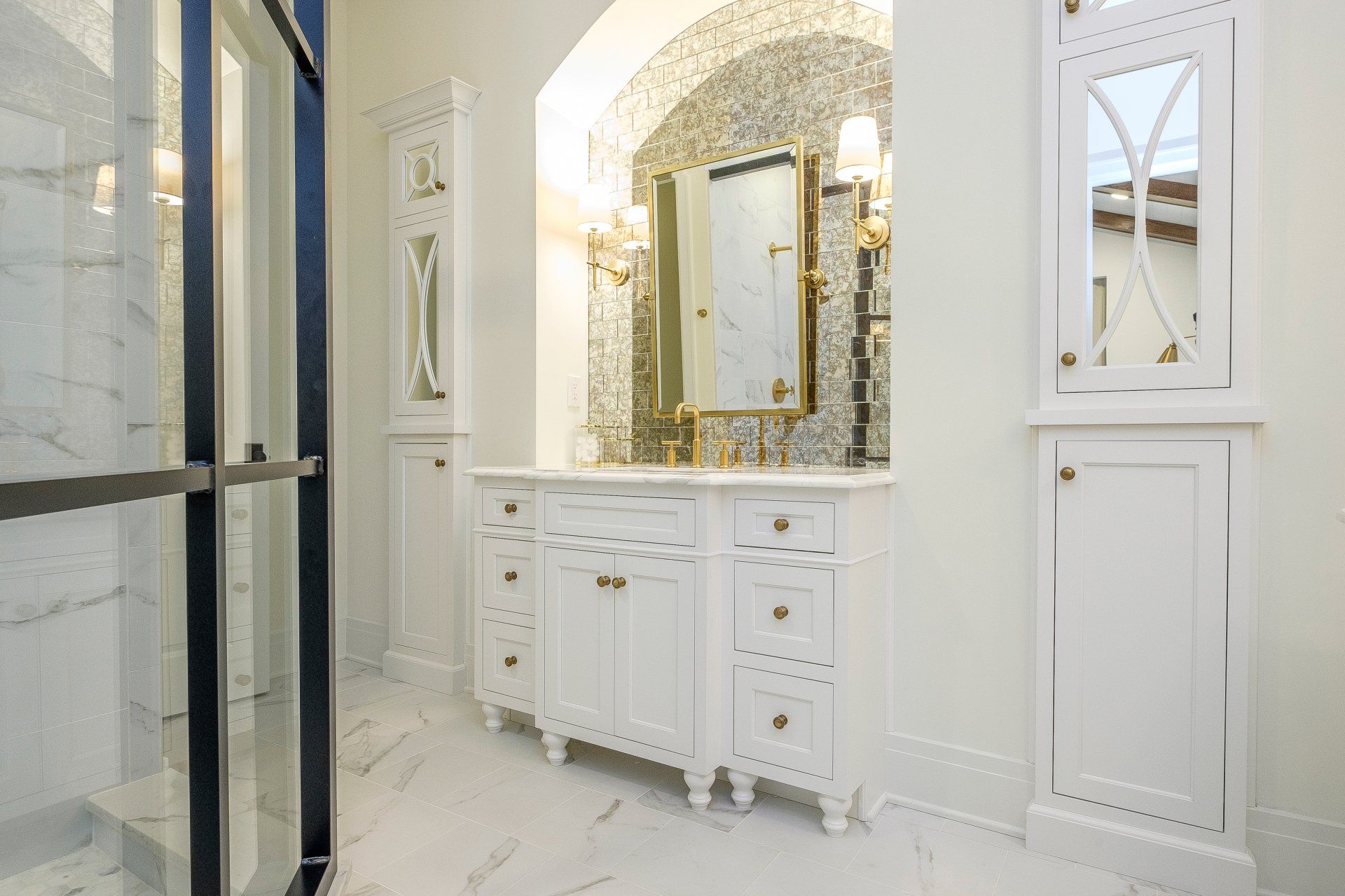 Elegant white bathroom vanity with gold-framed mirror, marble walls, and glass shower door