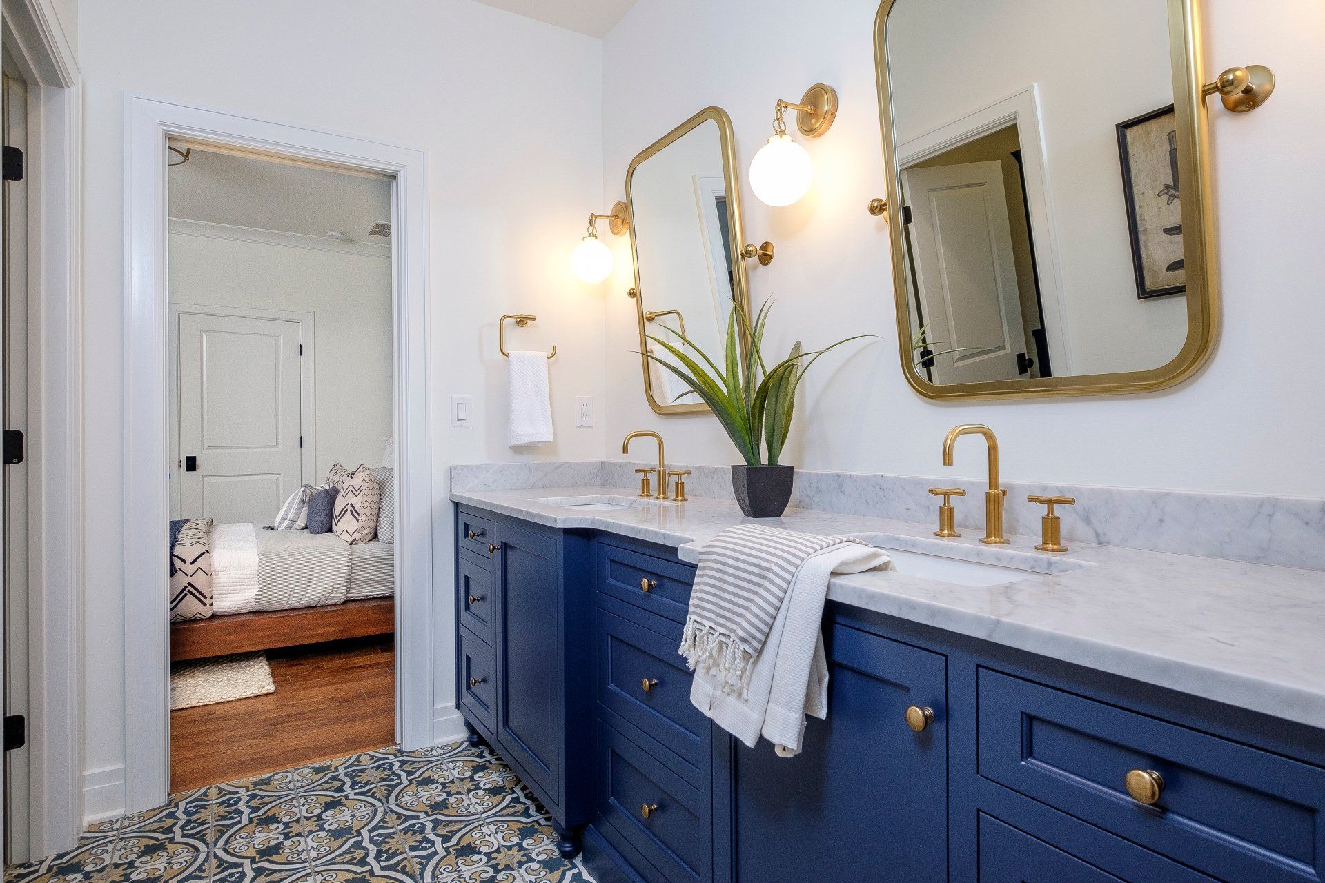 Blue bathroom vanity with gold fixtures, large mirror, and patterned floor tile; bedroom visible through open doorway