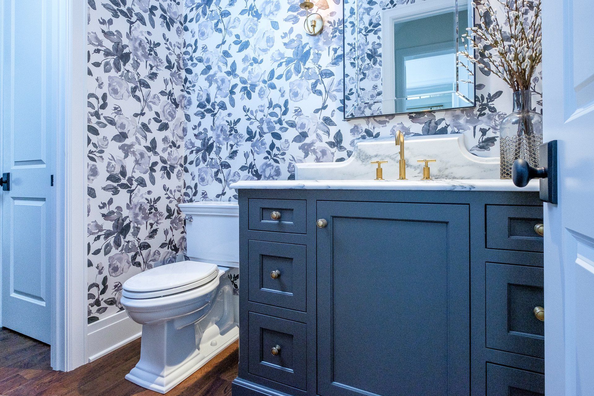 Small bathroom with floral wallpaper, blue vanity, white toilet, and gold faucet set under a mirror.