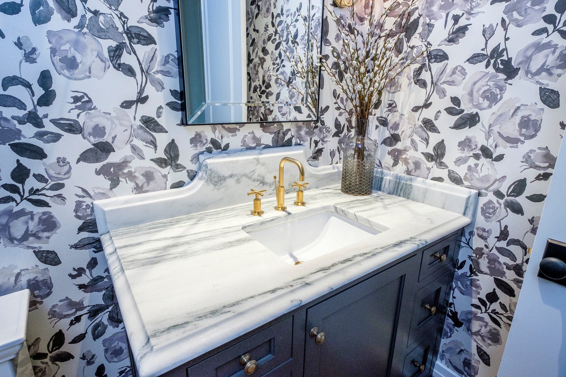 Bathroom vanity with white marble countertop, gold faucet, floral wallpaper, and a vase of dried branches