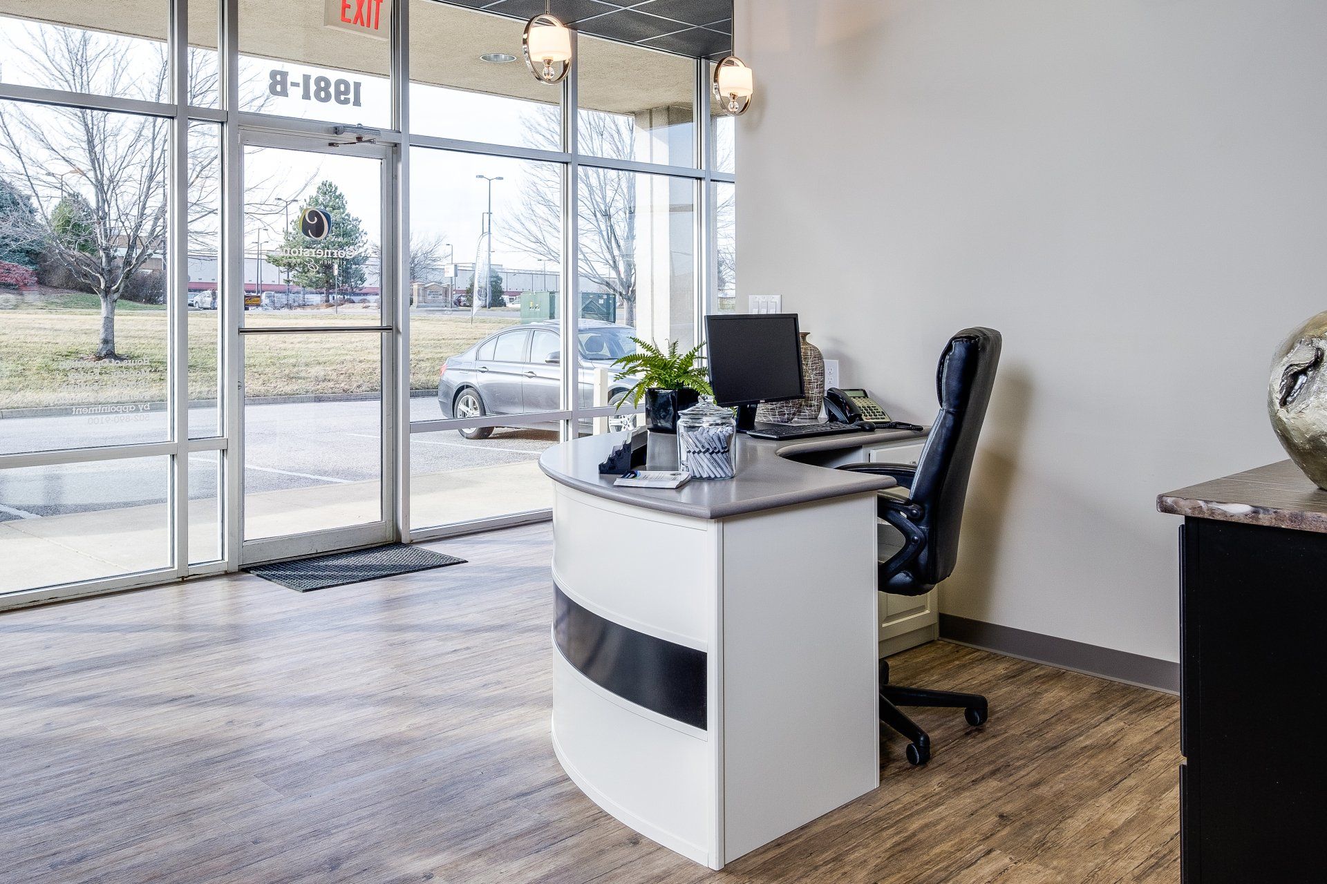 Office reception desk by glass entrance doors with a computer, chair, and potted plant.