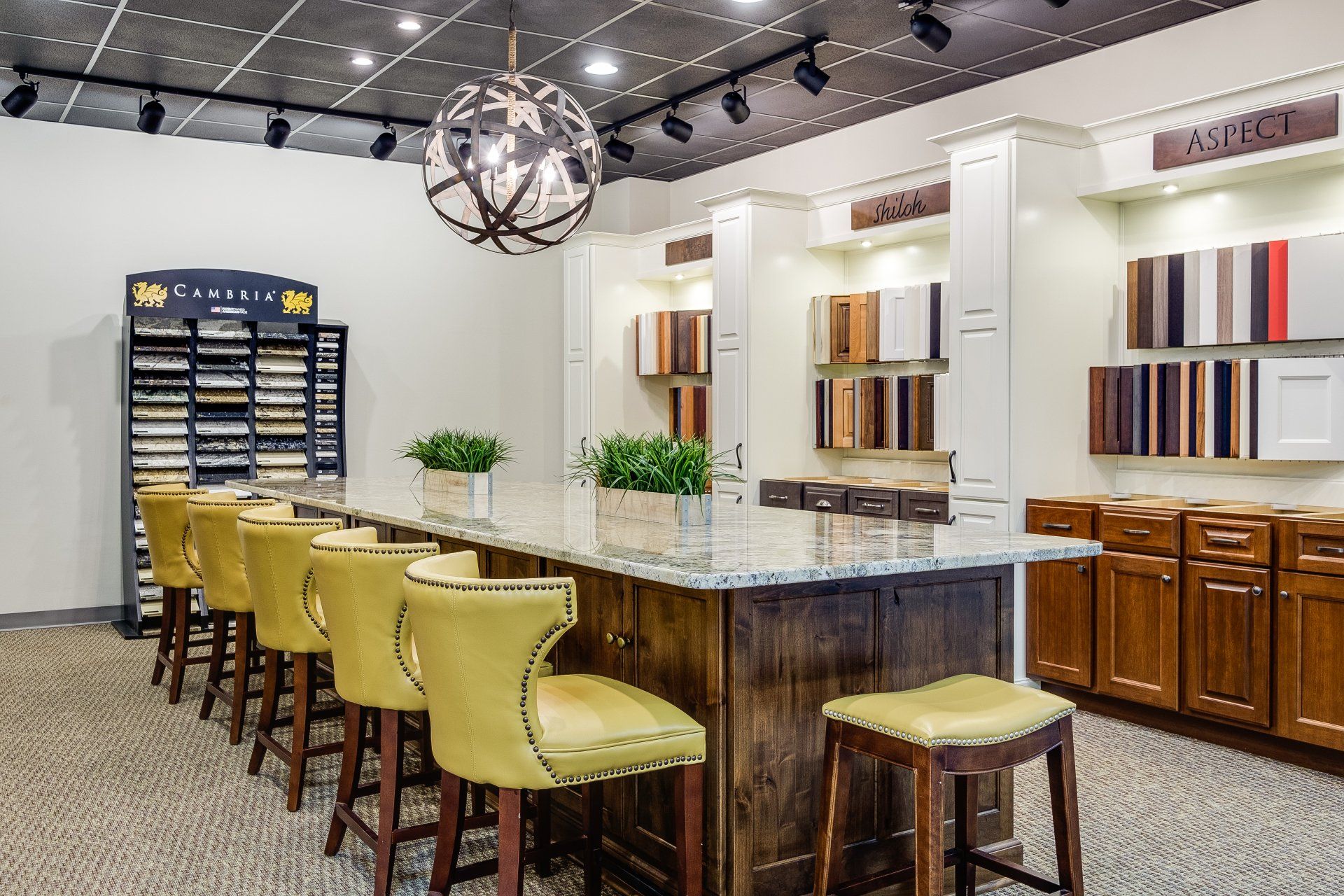 Bright showroom kitchen with granite island, yellow stools, cabinet displays, and hanging lights
