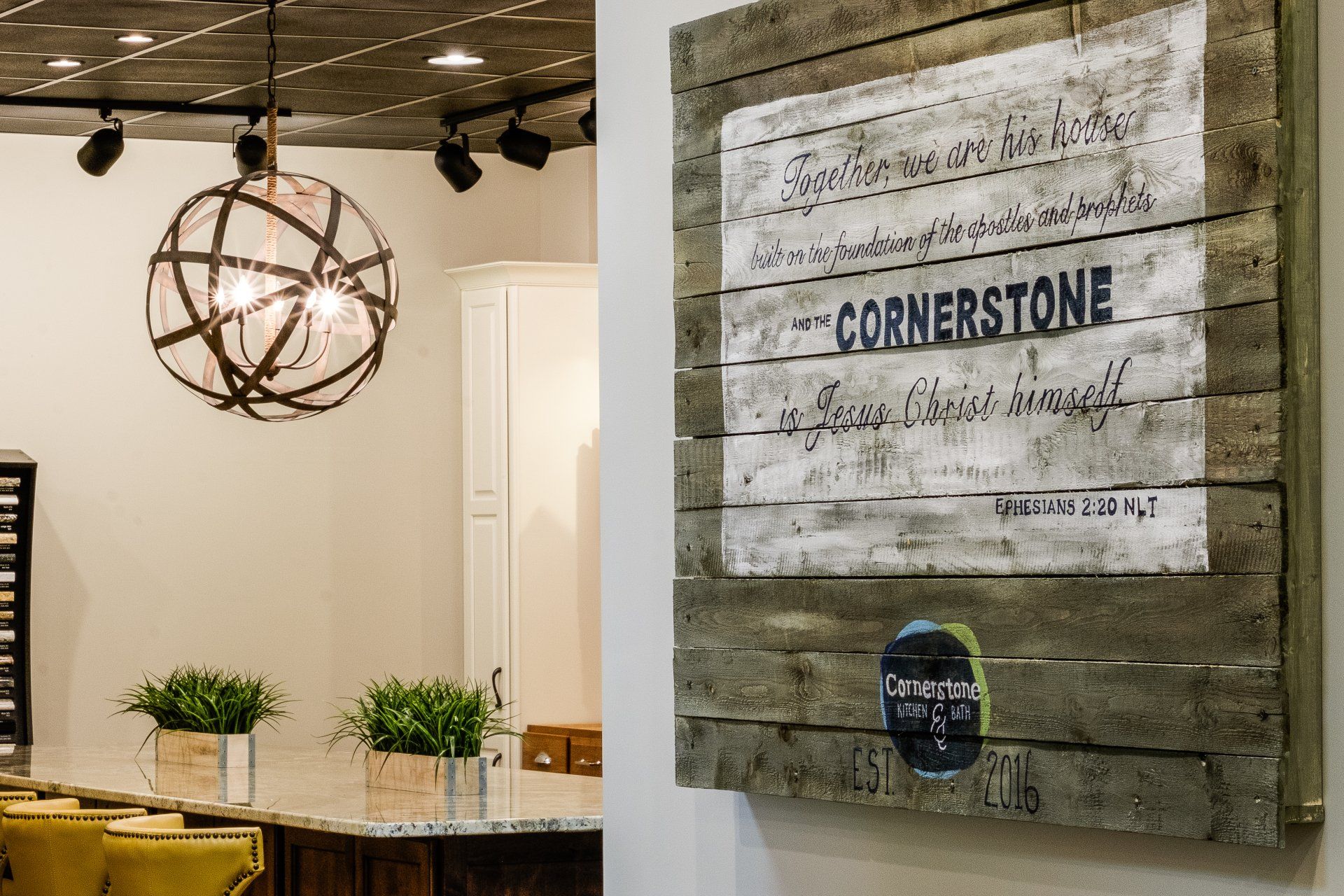 Office interior with hanging spherical light, wooden table, plants, and a wall sign reading “CORNERSTONE”