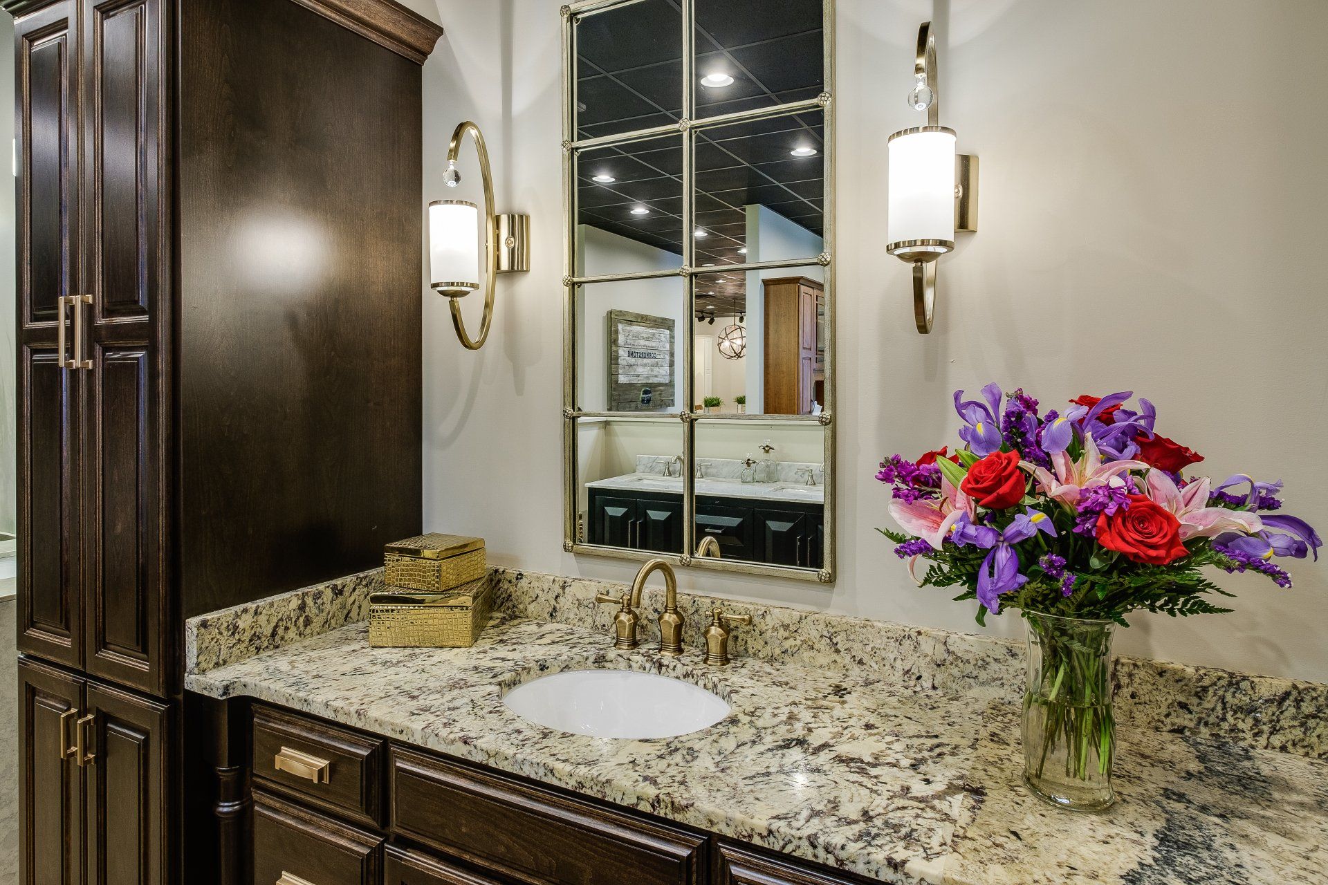 Bathroom vanity with granite countertop, mirror, wall sconces, and a vase of colorful flowers