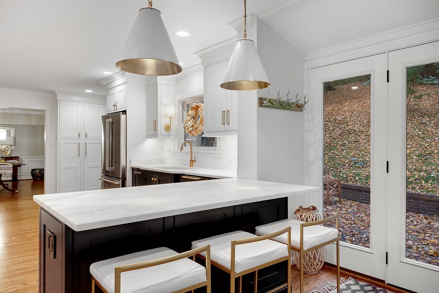 Bright modern kitchen with white island, pendant lights, gold stools, and glass doors to the right