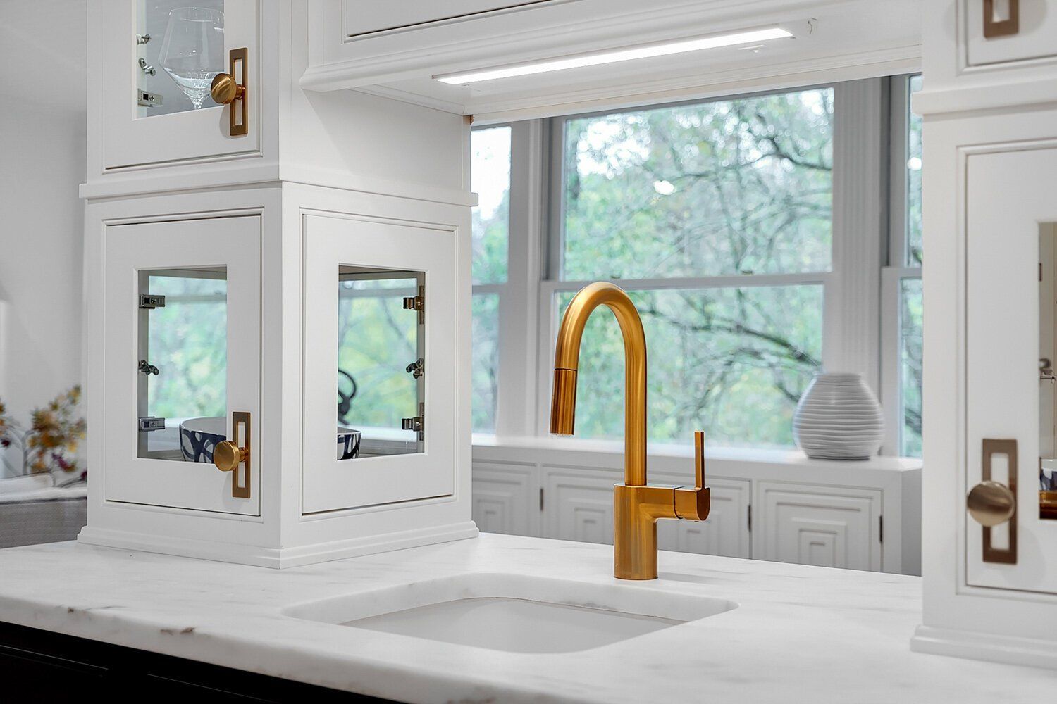 Bright white kitchen with a gold faucet, marble island, and large window looking onto trees