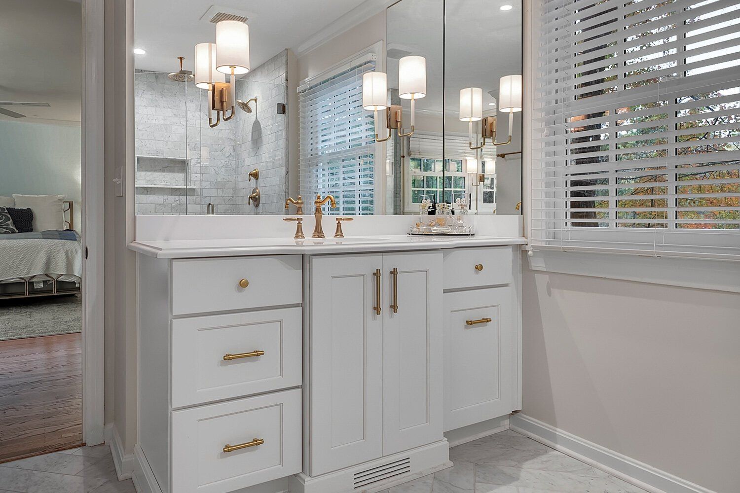 Bright white bathroom vanity with gold hardware, large mirror, wall sconces, and window blinds