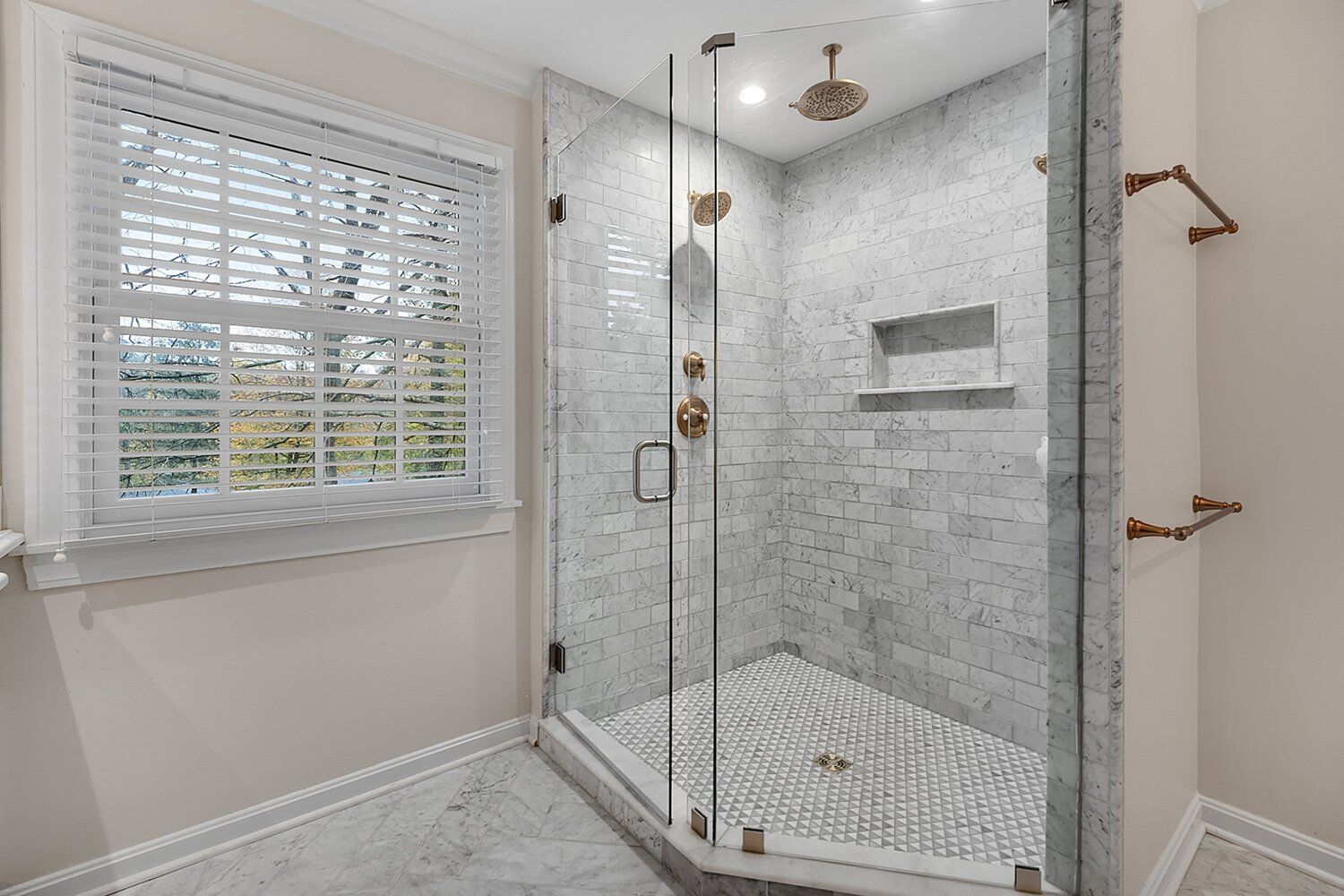 Bright bathroom with a glass walk-in shower, marble tile, and a window with white blinds.