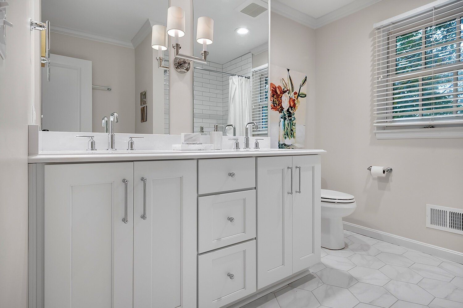 Bright white bathroom with double vanity, large mirror, chandelier, and window blinds.