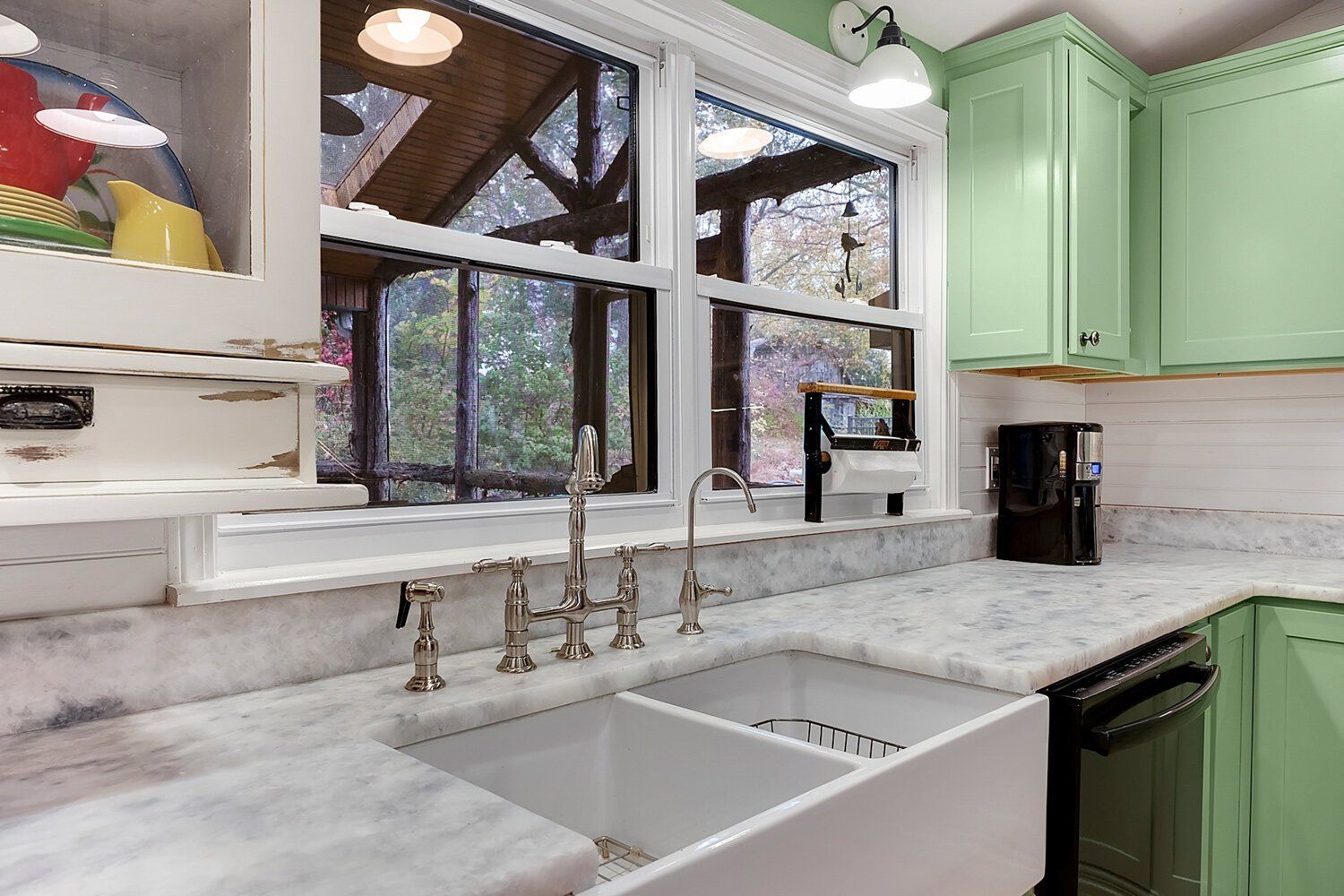 Bright kitchen with green cabinets, marble counters, white sink, and large windows overlooking trees
