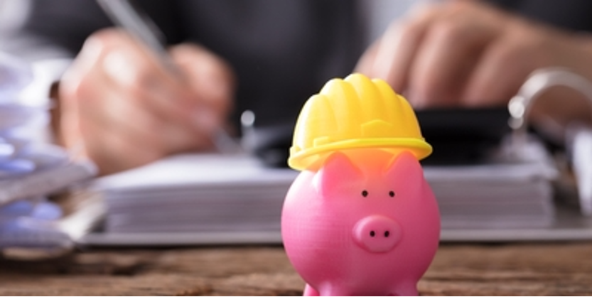 Pink piggy bank wearing a yellow hard hat on a desk with blurred hands in the background
