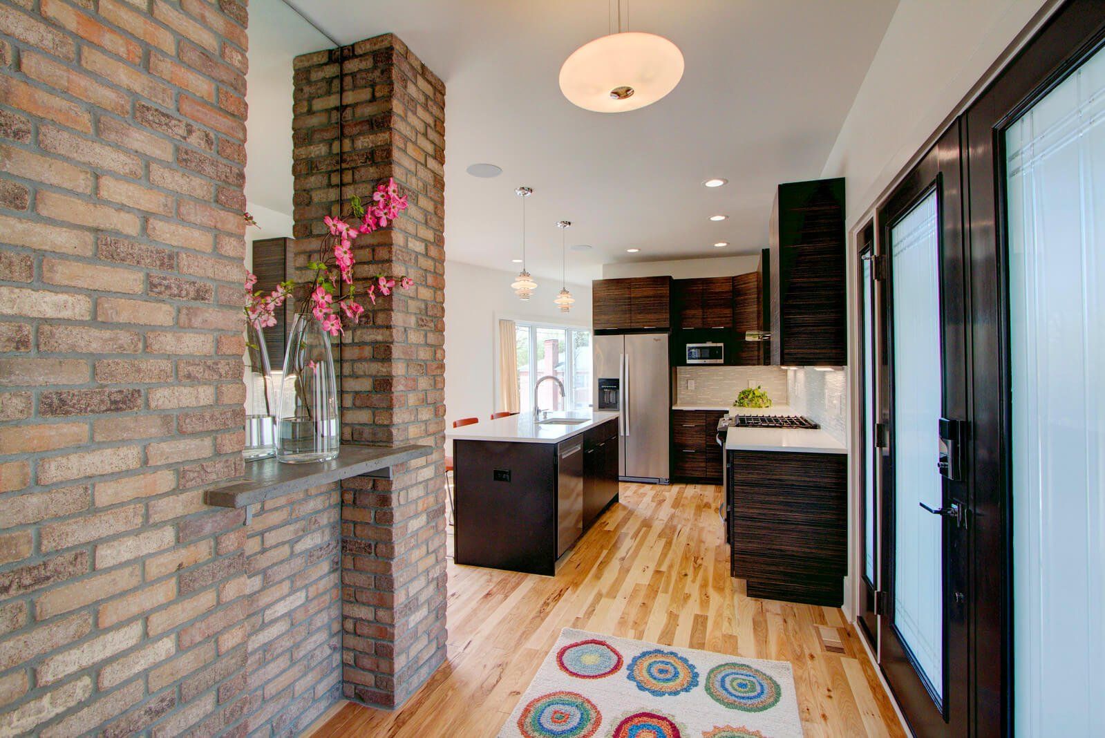 Modern kitchen with brick walls, dark cabinets, wood floor, and colorful rug in a bright hallway