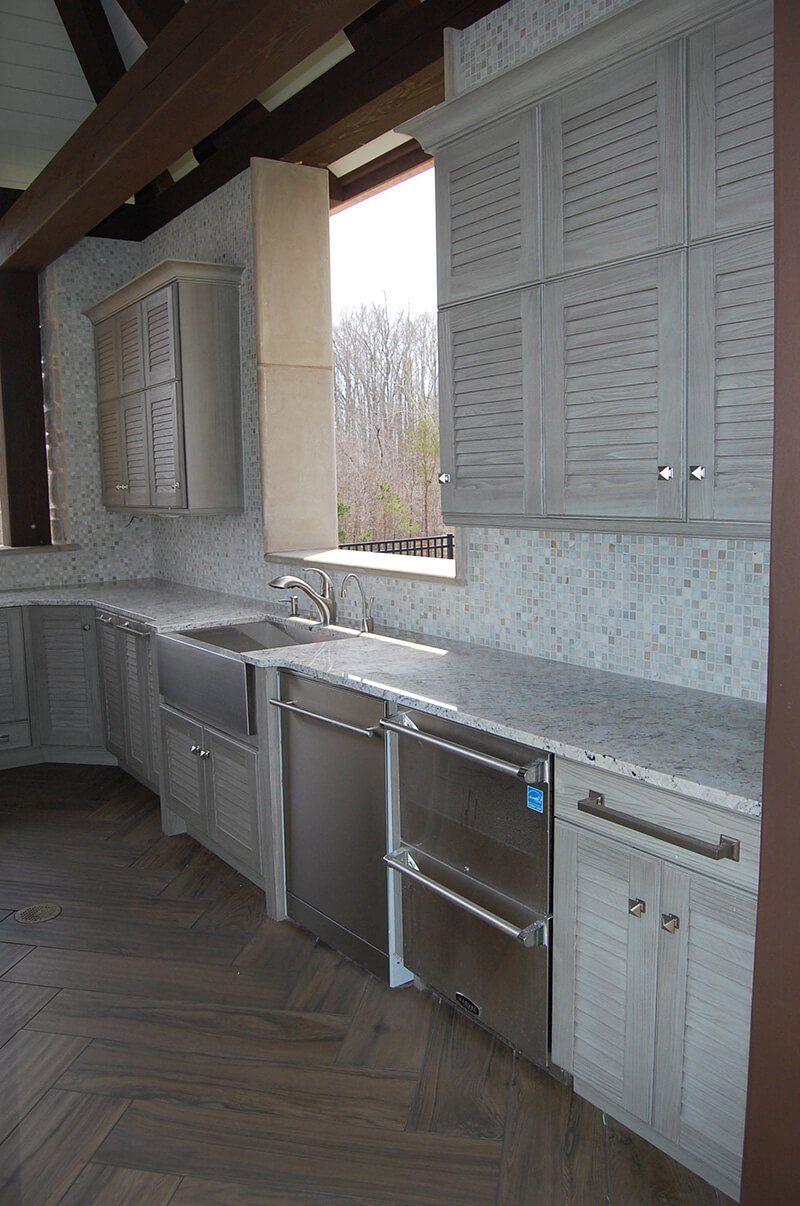 Gray outdoor kitchen with cabinets, sink, and countertop under a sloped roof, overlooking trees