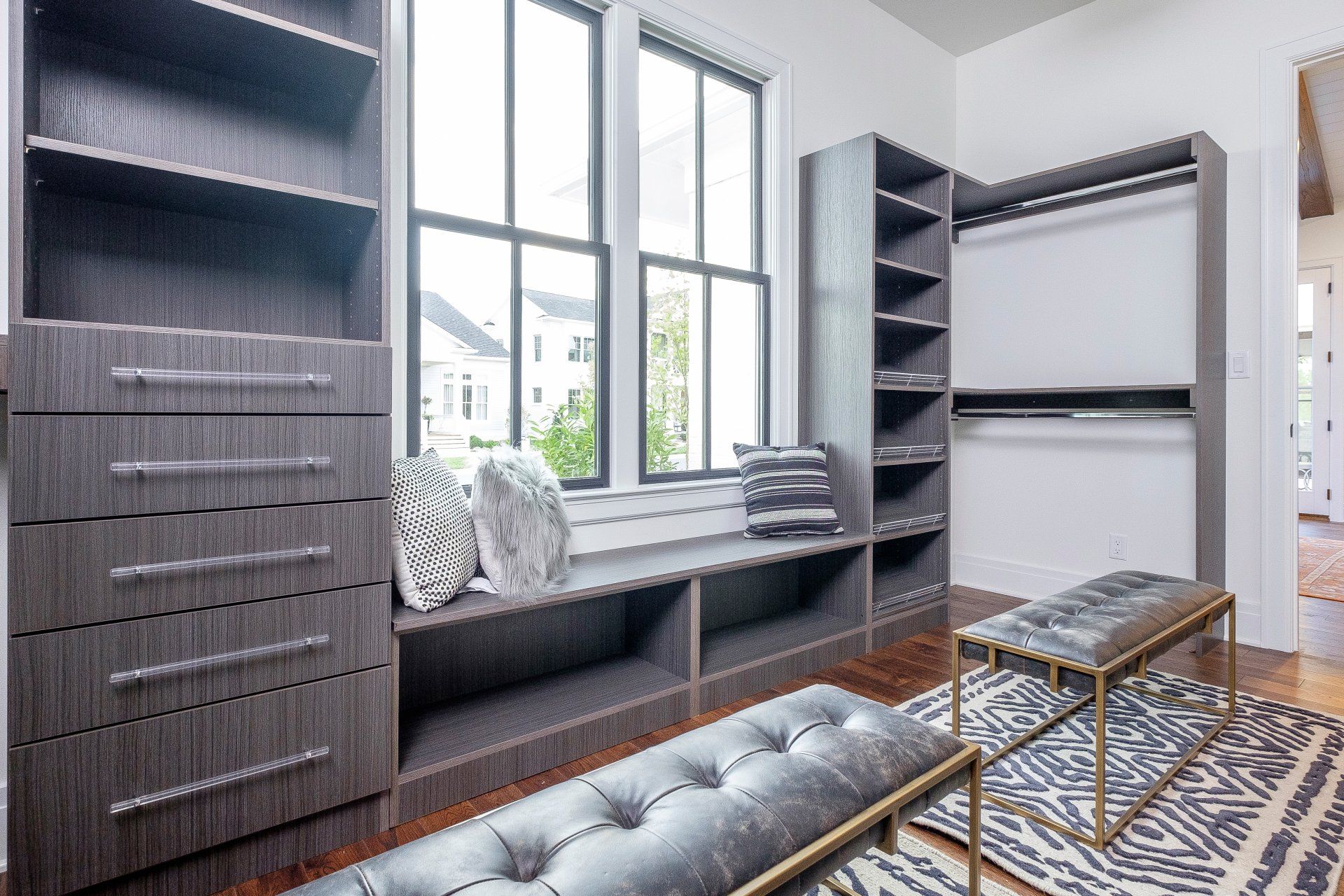 Modern walk-in closet with dark built-in shelves, drawers, bench seating, and a patterned rug by a window.