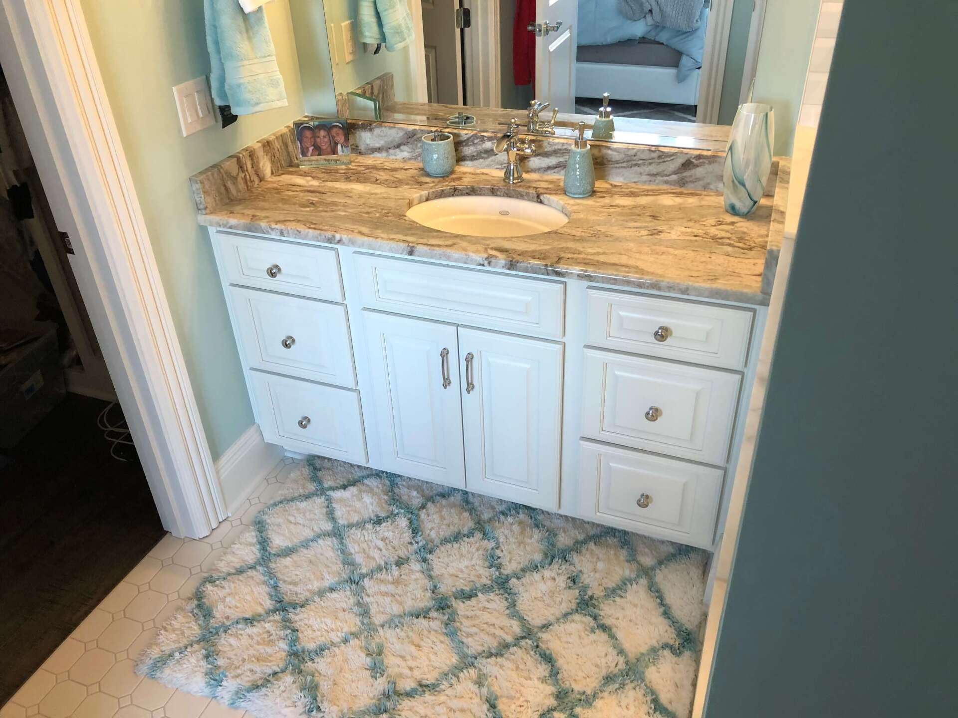 Bathroom vanity with white cabinets, marble countertop, and patterned rug in front of a mirror
