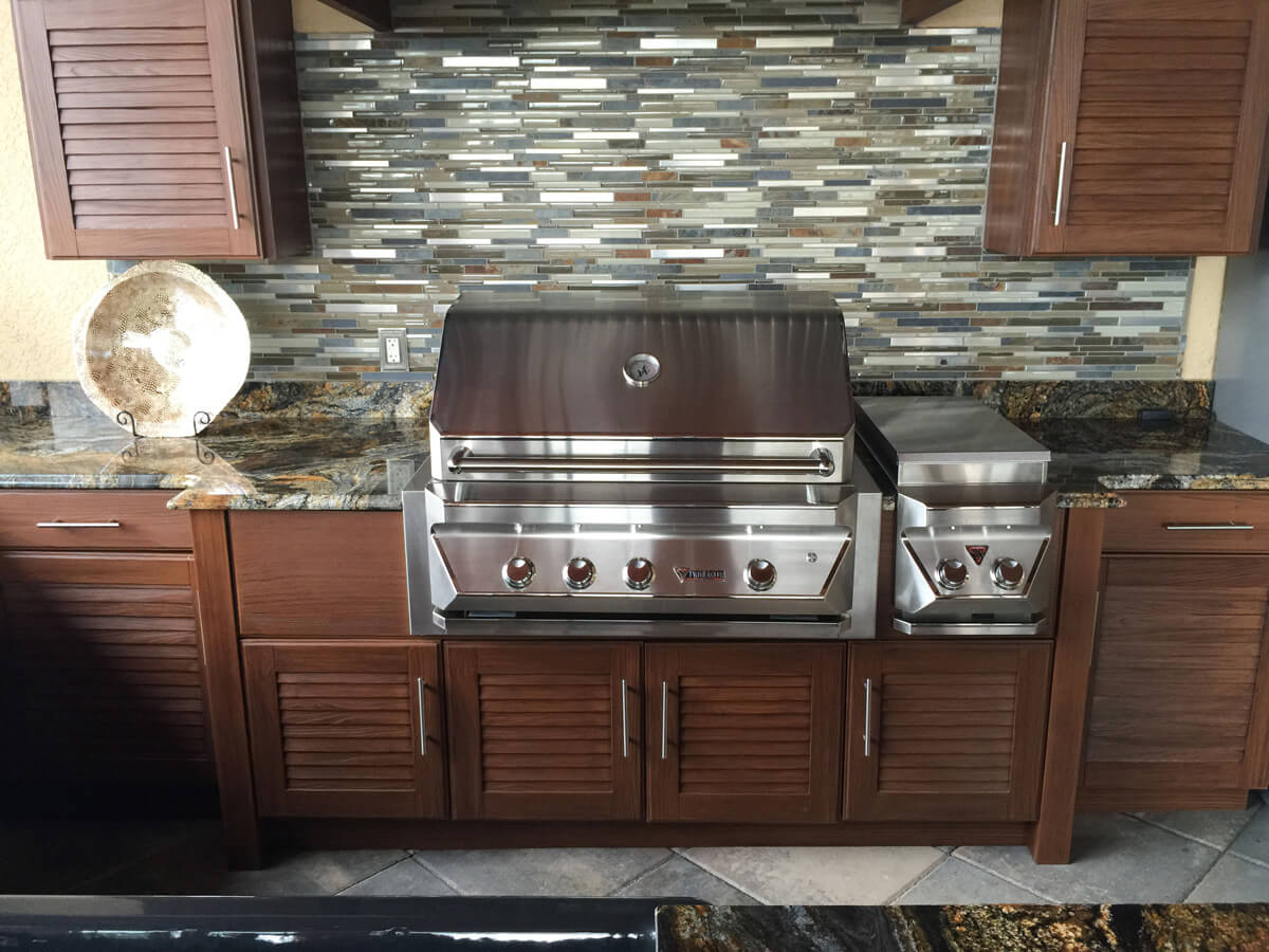 Outdoor kitchen with stainless steel grill, granite counters, and dark wood cabinets against a stone backsplash