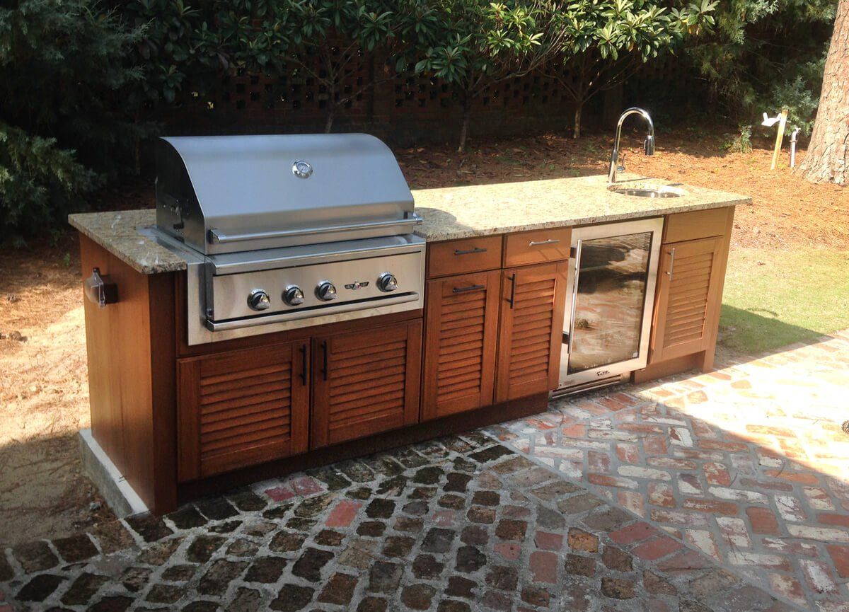 Outdoor stainless-steel gas grill and wooden cabinet island on a brick patio, beside a sink.