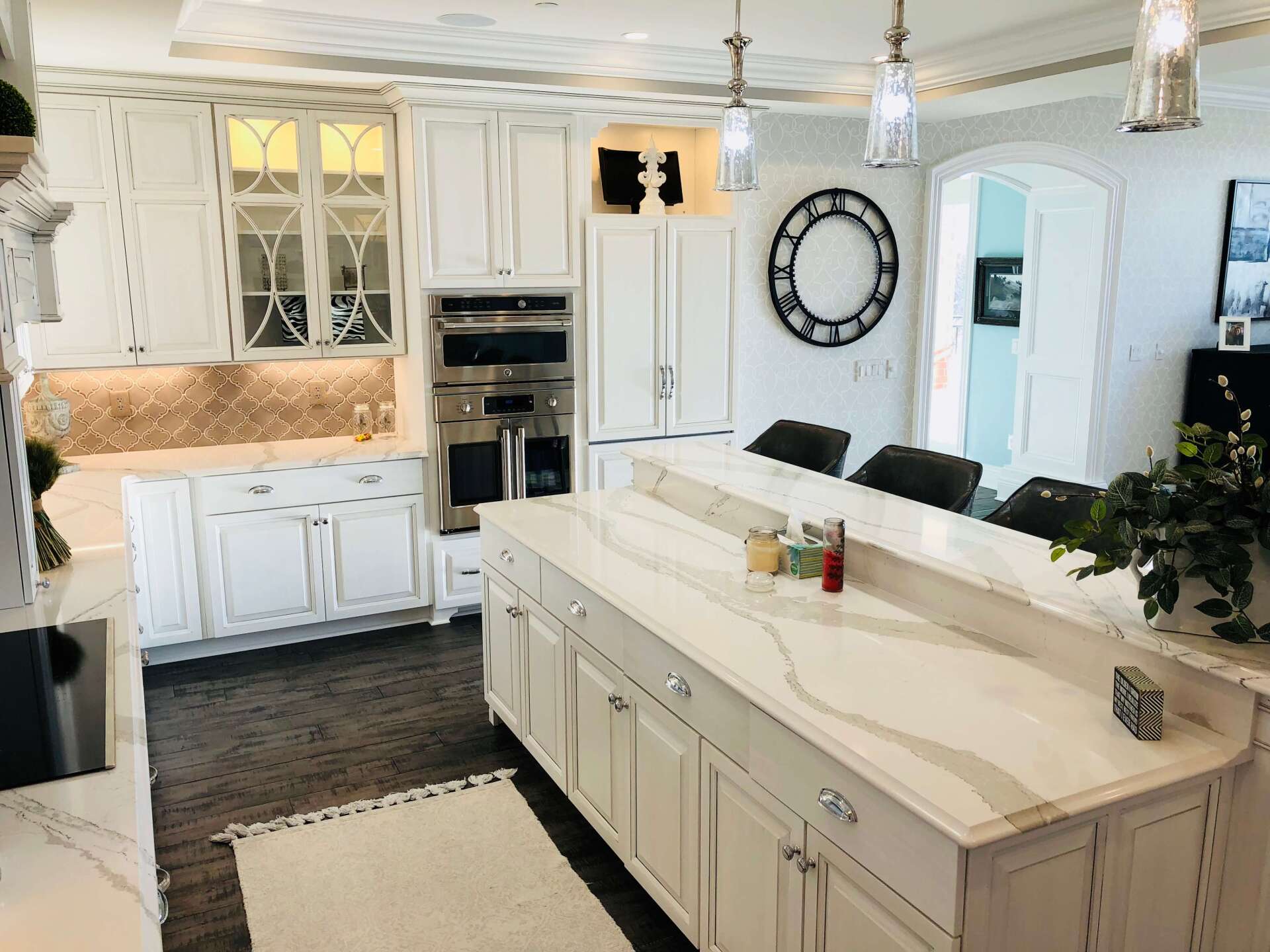 Bright white kitchen with large island, built-in ovens, dark wood floors, and hanging pendant lights.