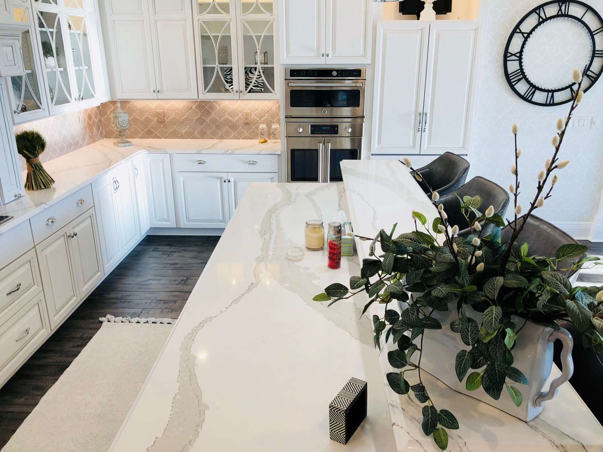 Bright white kitchen with marble island, built-in ovens, and large wall clock