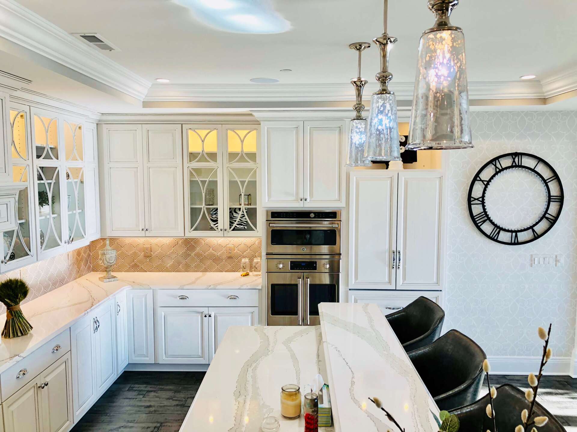 Bright white kitchen with marble counters, stainless appliances, pendant lights, and a large wall clock.