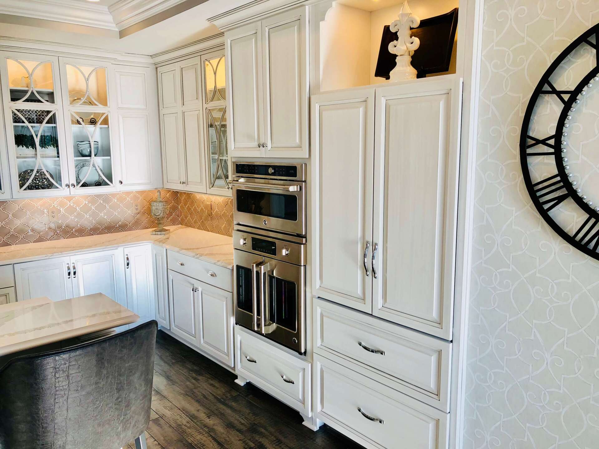 Bright white kitchen with marble countertops, built-in ovens, cabinets, and a large black wall clock