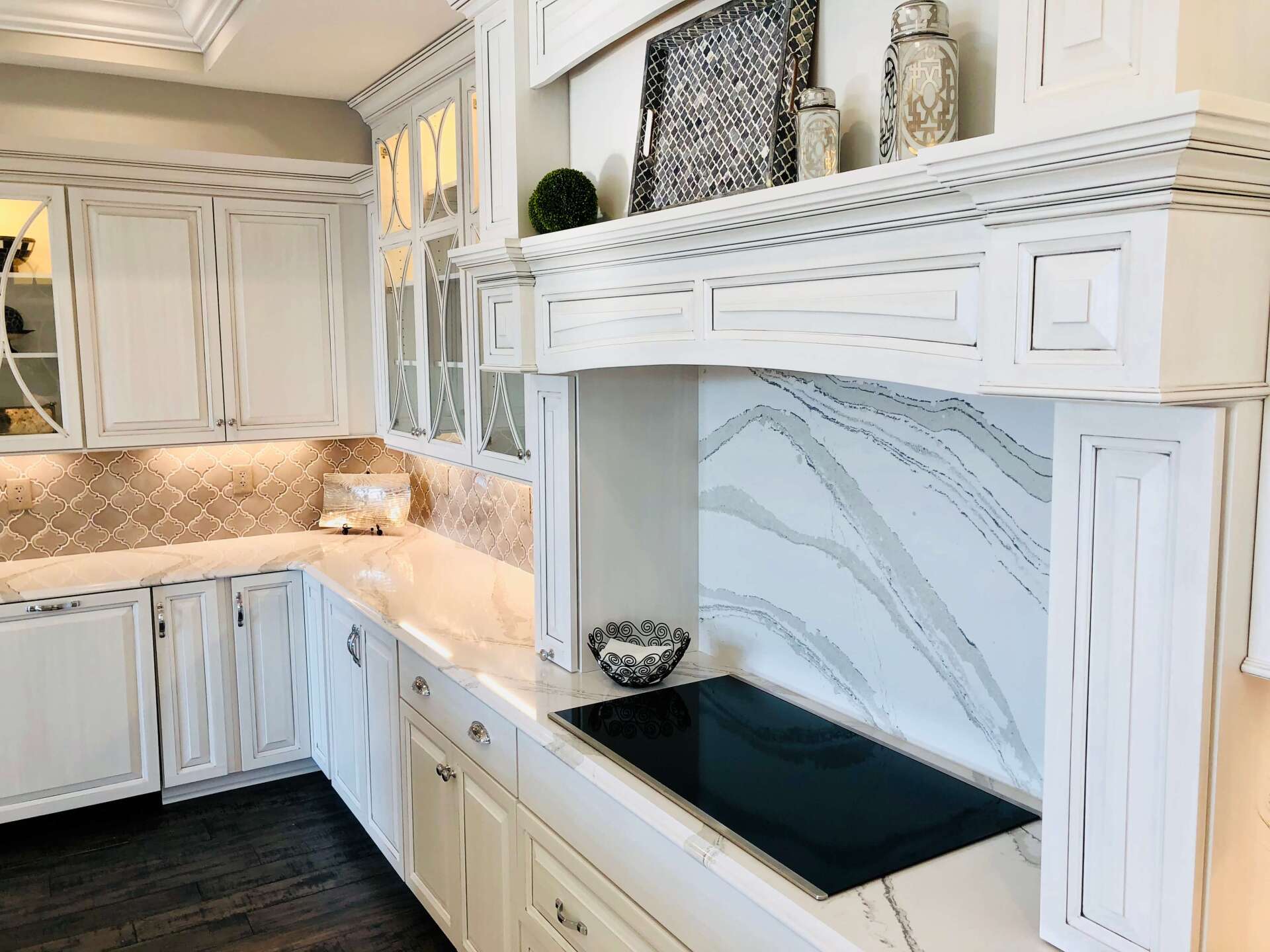White kitchen with marble countertops, ornate cabinets, and a black cooktop beneath a decorative range hood