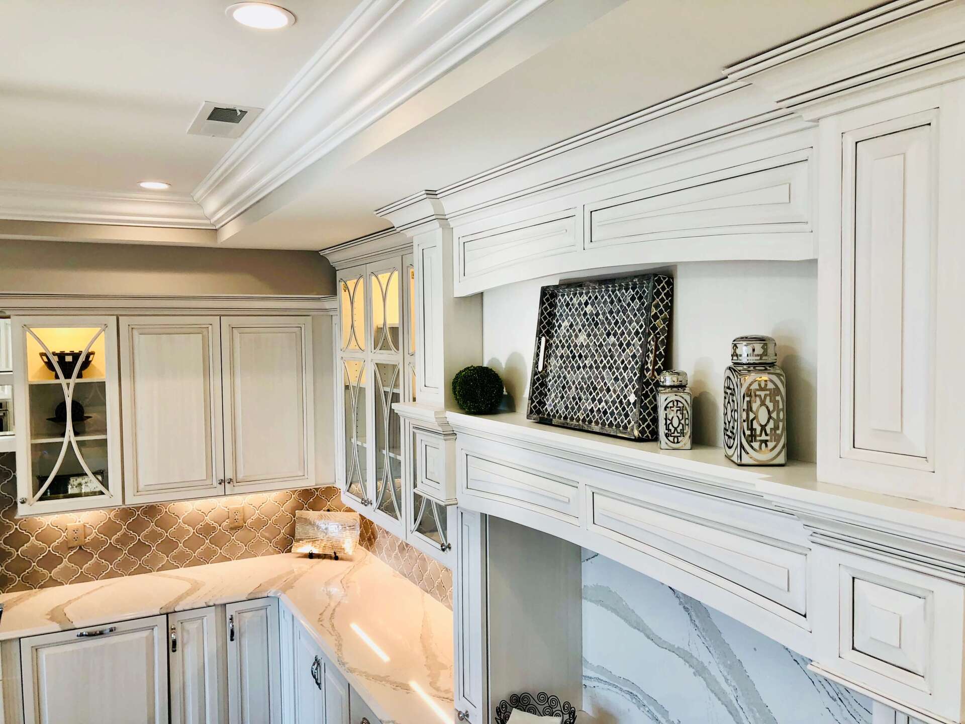 Bright white kitchen with ornate cabinets, marble countertop, and decorative shelves with vases.
