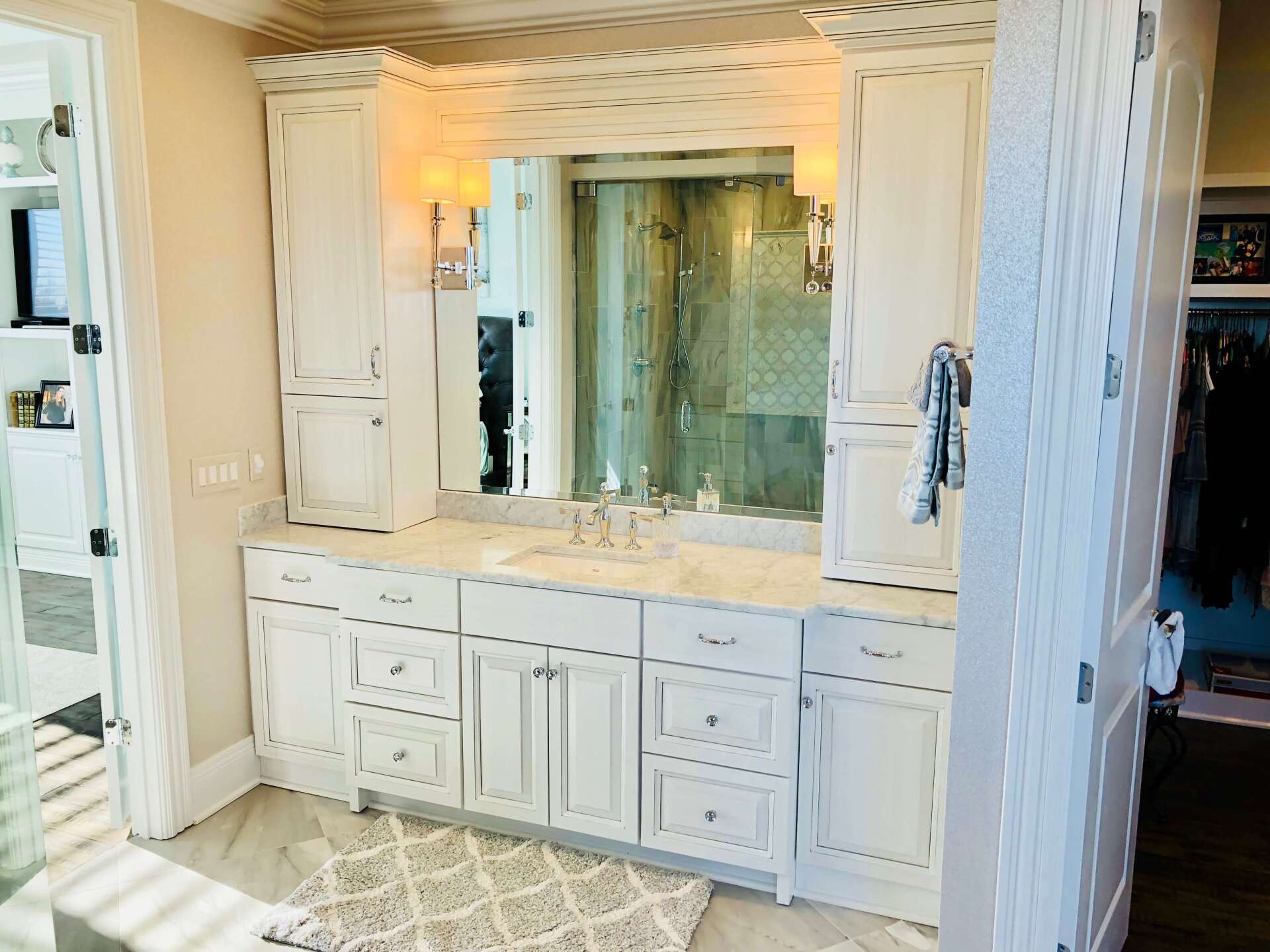 White bathroom vanity with large mirror, sink, cabinets, and a lit wall sconce in a bright tiled room