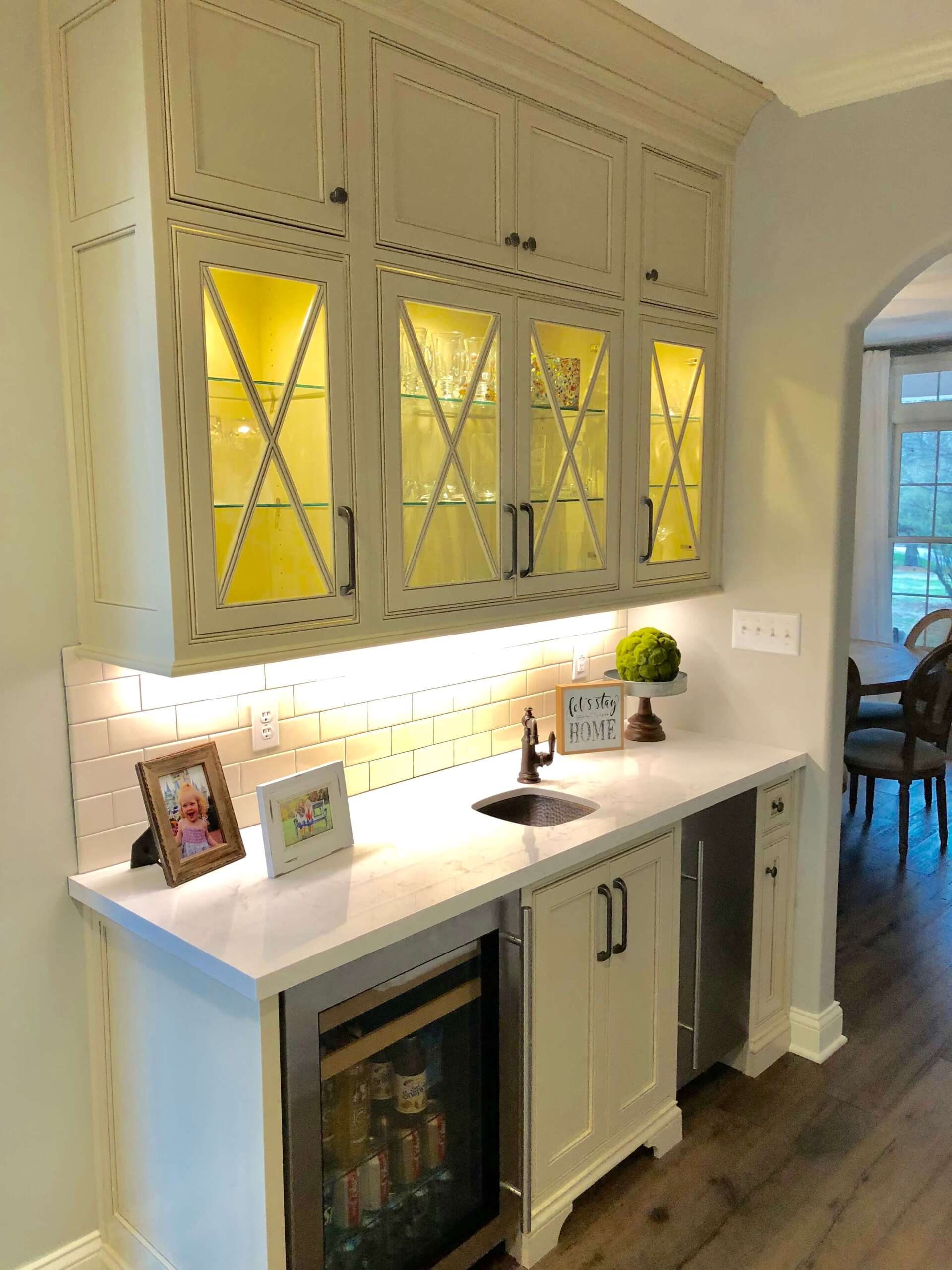 Cream kitchen with lit glass-front cabinets, white countertop, sink, and under-counter wine fridge.