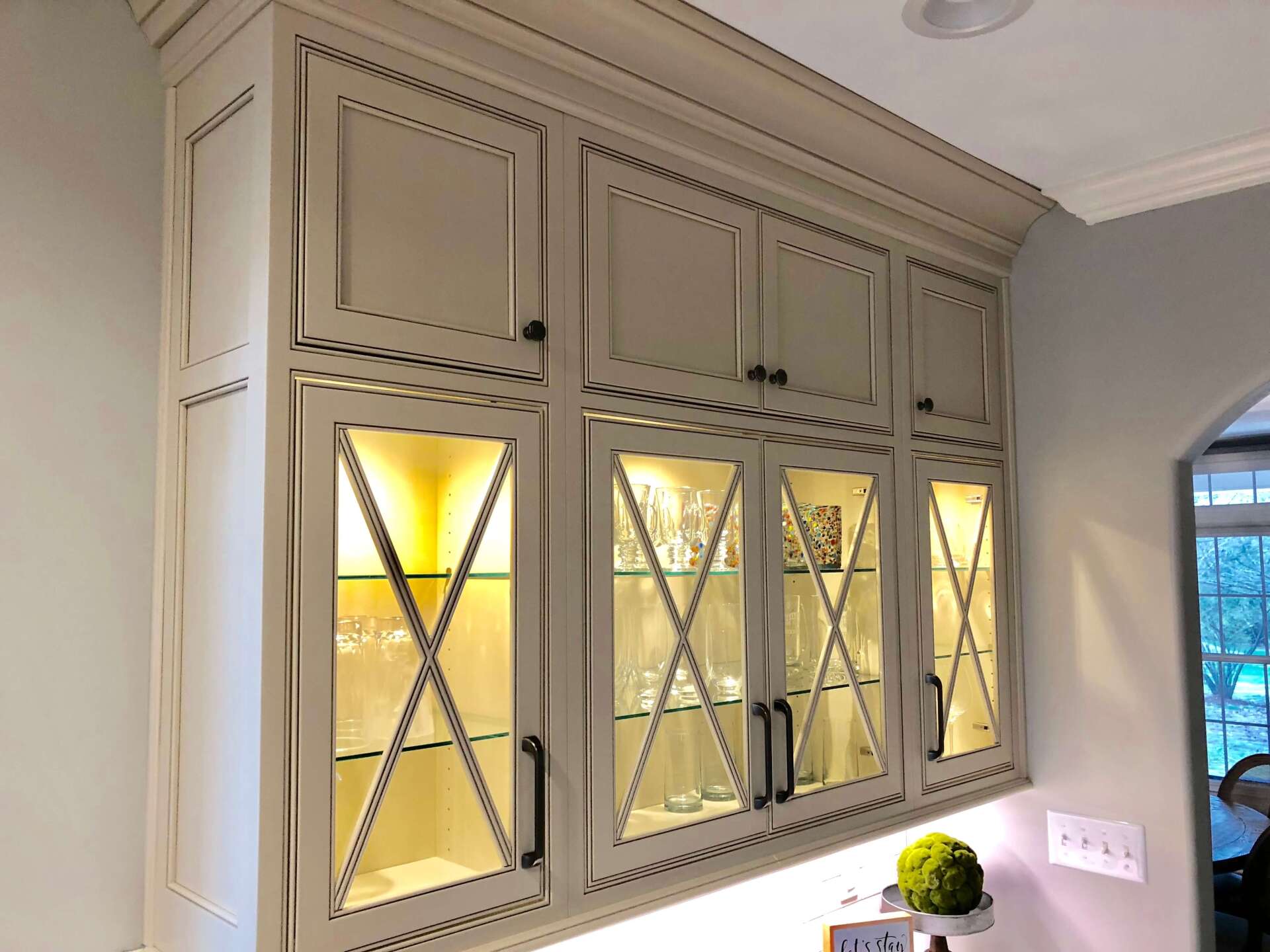 Built-in kitchen cabinets with glass doors and warm interior lighting against a gray wall