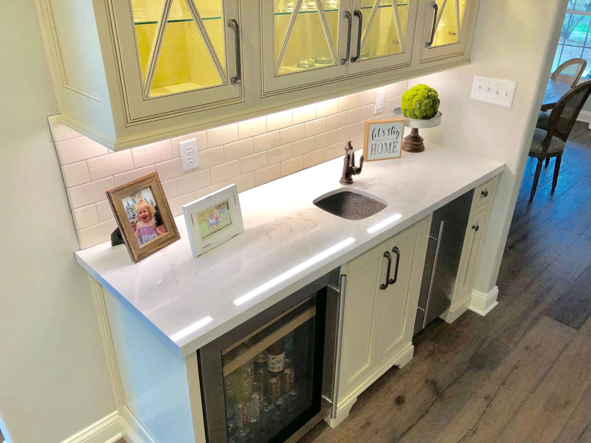 Small home bar with white countertop, glass-front wine fridge, sink, and decor under a mirrored cabinet.