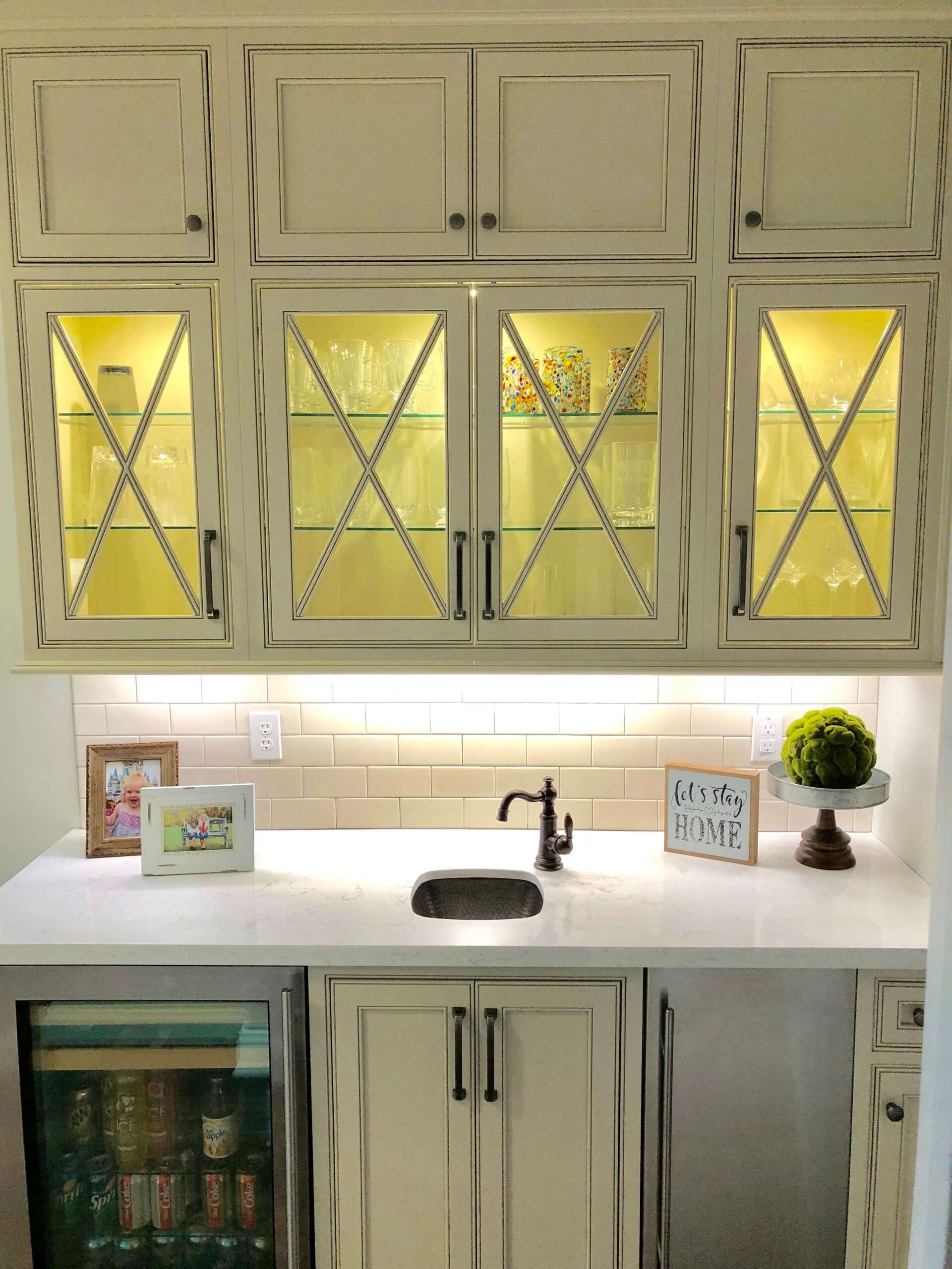 Bright kitchen with cream cabinets, white countertop, black sink, and warm yellow-lit window behind upper doors
