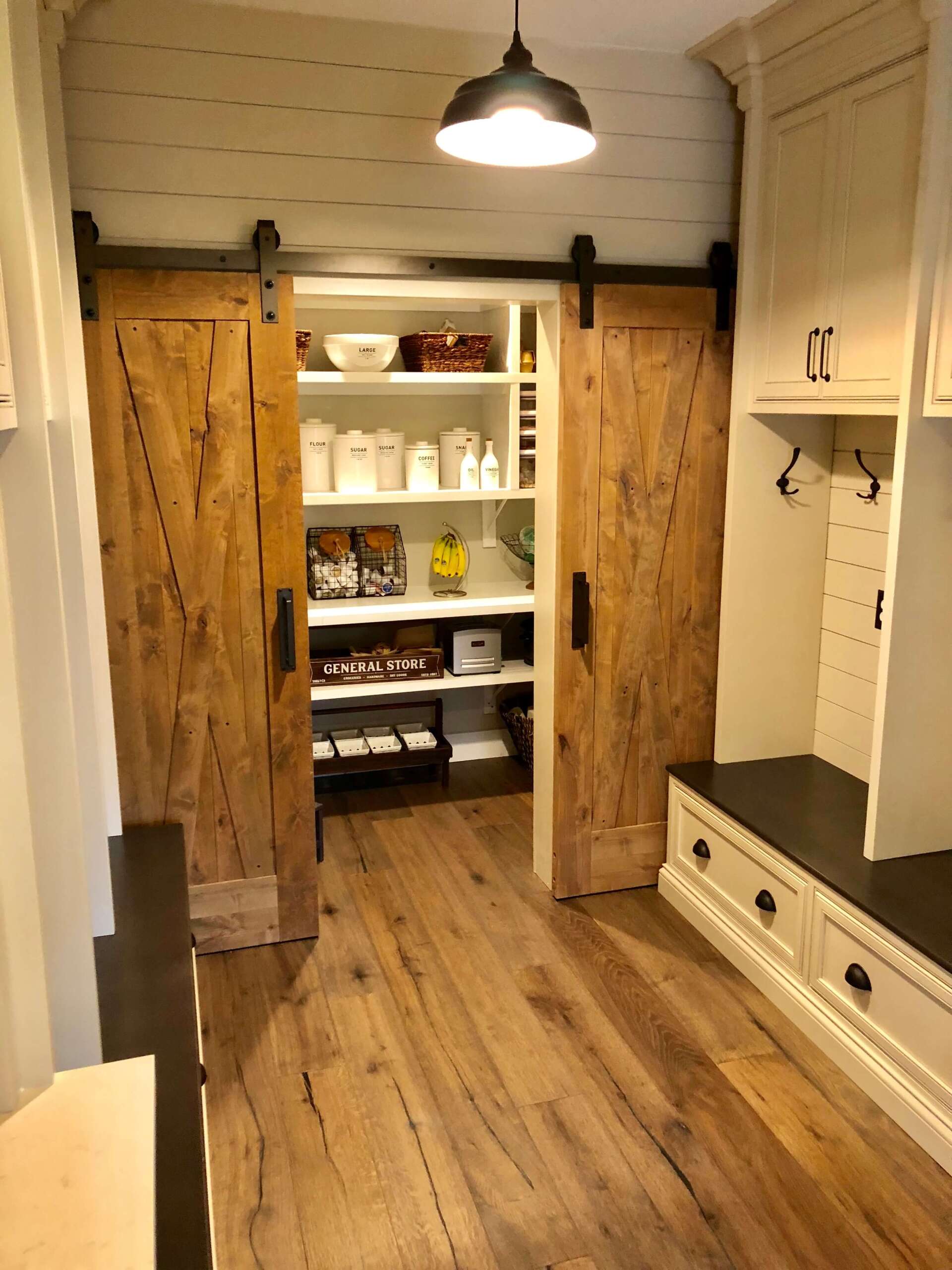 Rustic pantry with open wooden doors, white shelves, and jars in a bright hallway with wood floors.