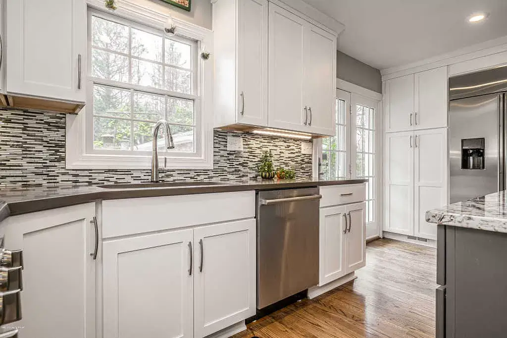 Bright white kitchen with mosaic backsplash, stainless dishwasher, and sunlit window over sink.