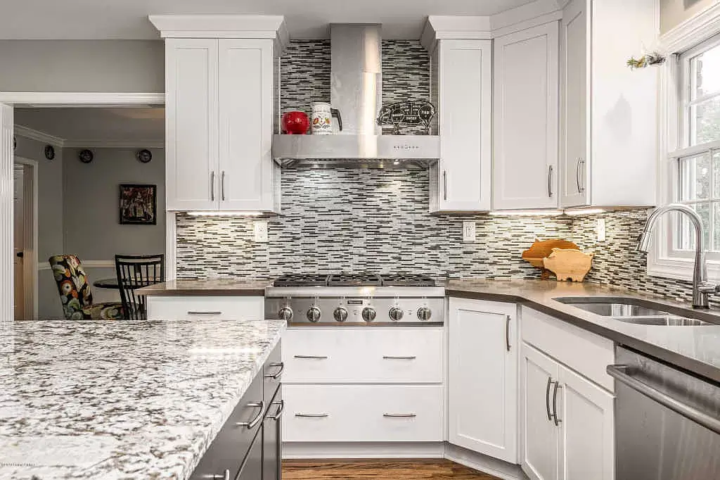 Bright white kitchen with granite countertops, mosaic backsplash, stainless stove, and a window by the sink