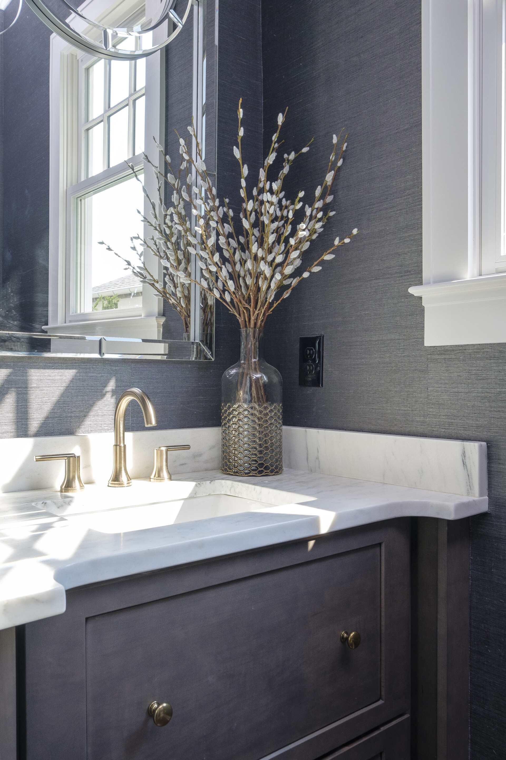 Modern bathroom vanity with white sink, gold faucet, dark cabinet, and vase of branches by a window