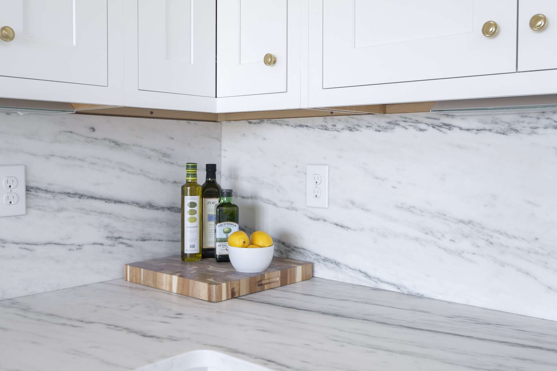 White kitchen counter with olive oil, lemons, and jars on a wooden tray against a marble backsplash